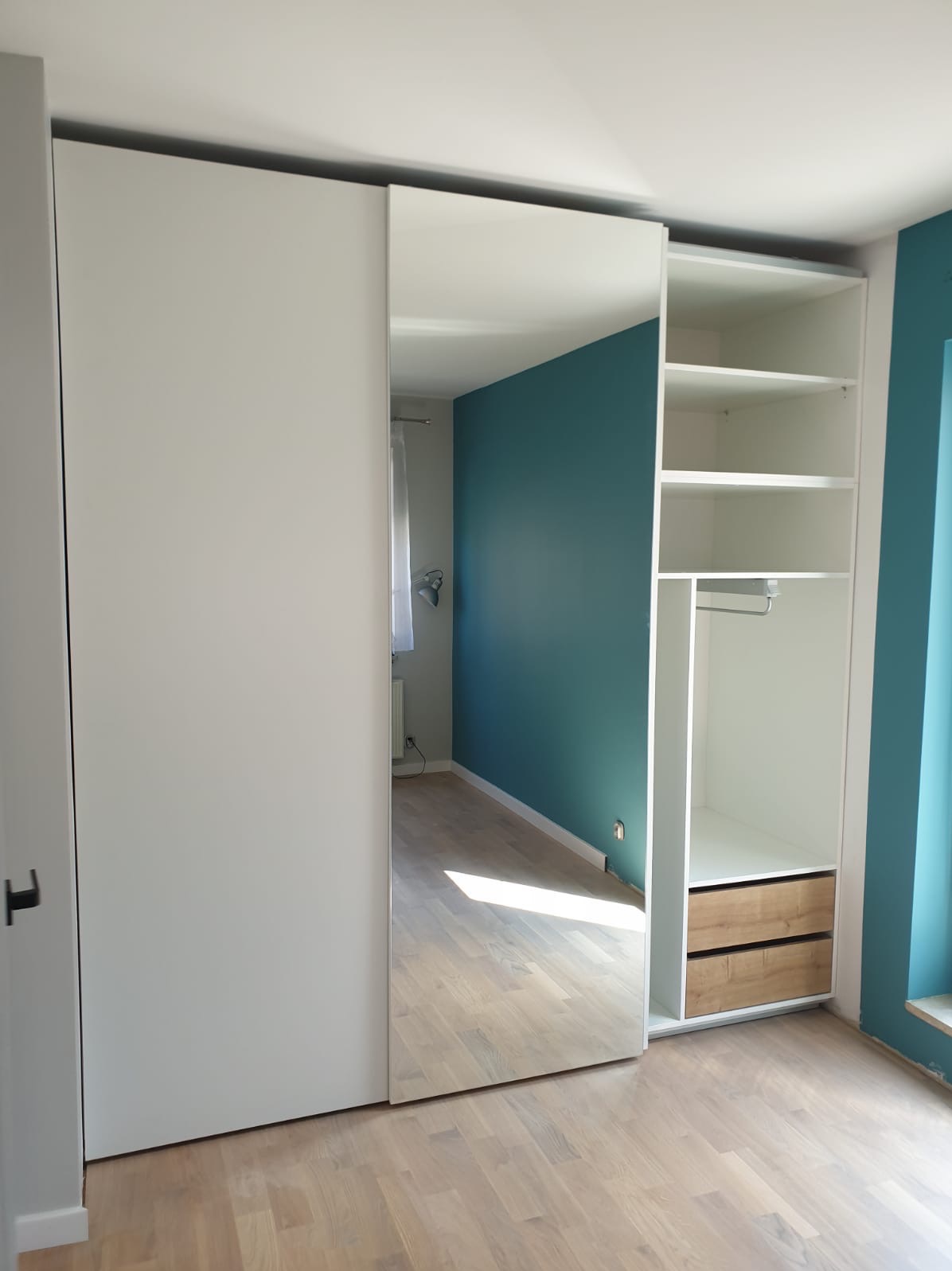 Biała szafa z przesuwnymi drzwiami z lustrem, widoczne półki i dwie szuflady z jasnego drewna, na podłodze panele w odcieniach beżu, ściana w kolorze turkusowym.
