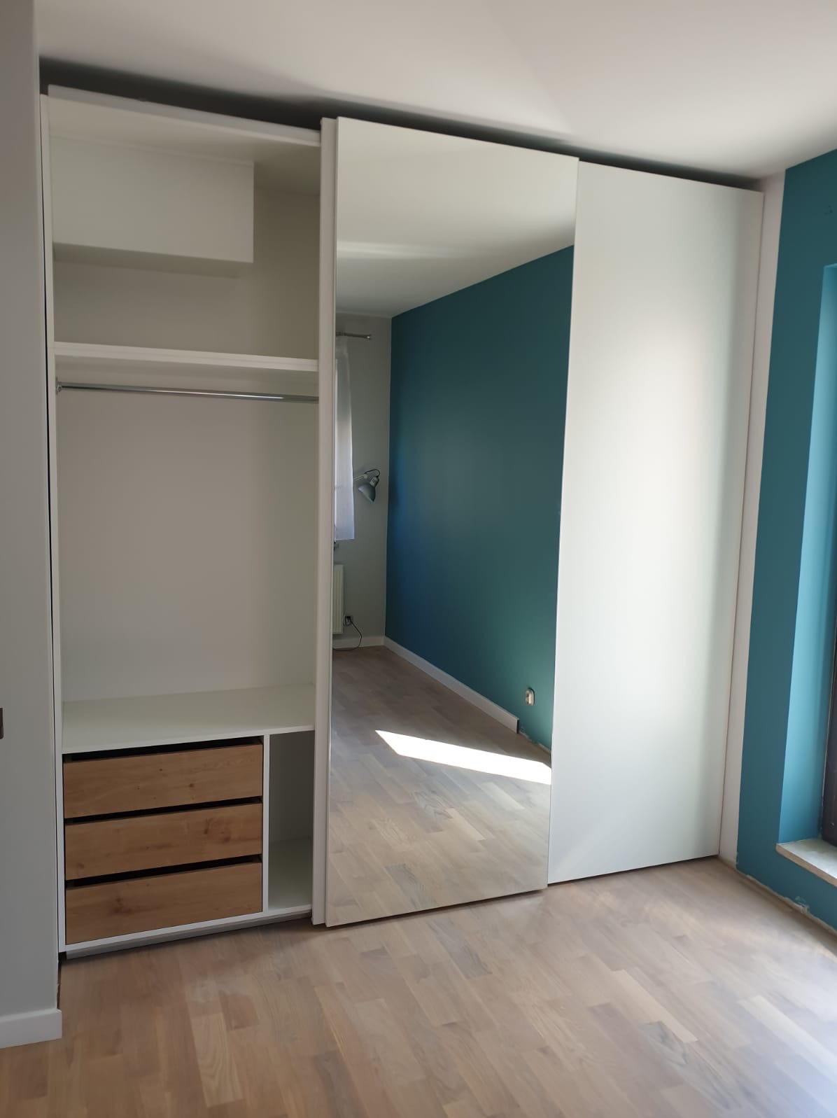 Biała szafa przesuwna z lustrem i drewnianymi szufladami, widok na wnętrze szafy i odbicie pokoju z turkusową ścianą.