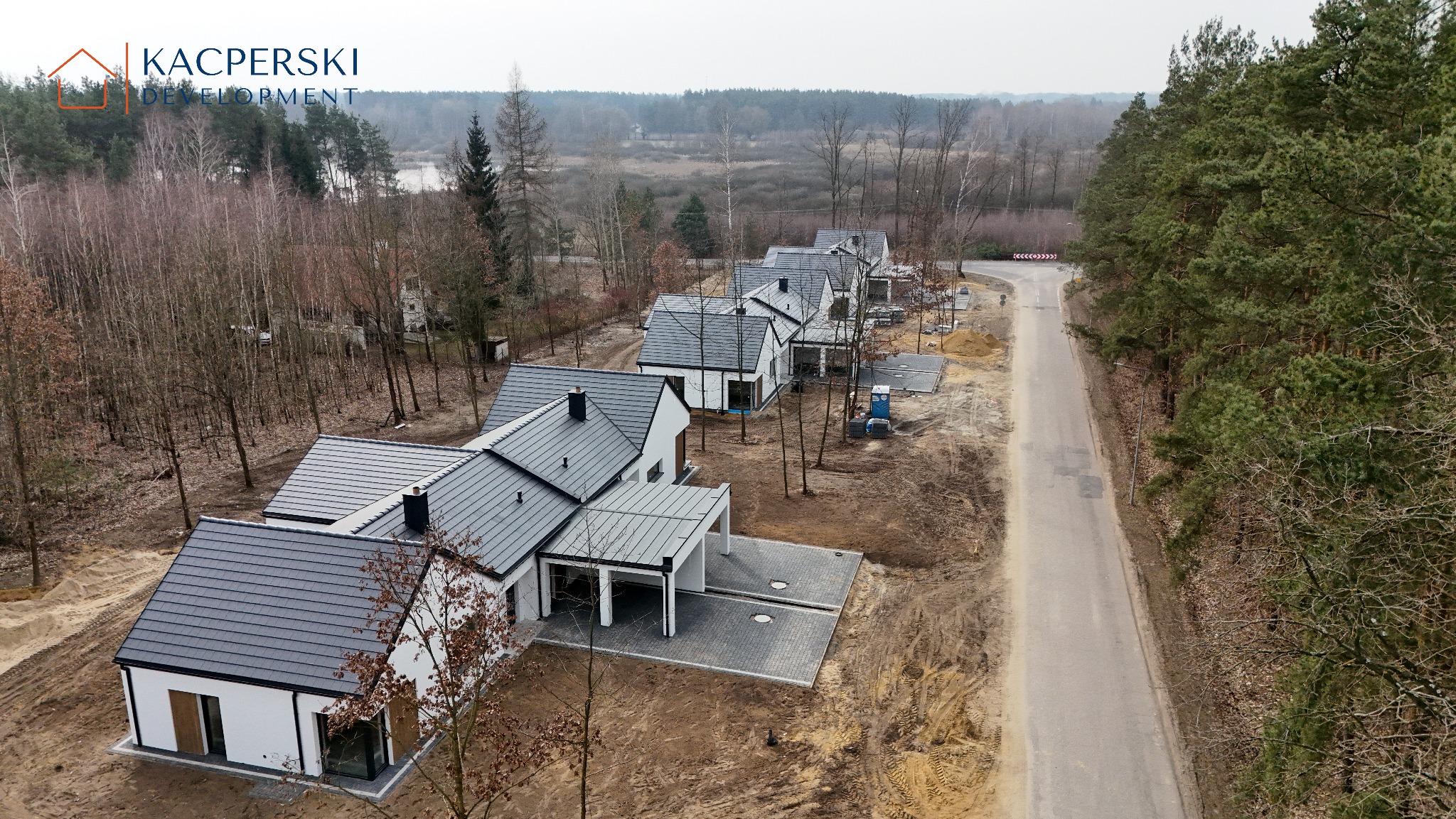 Osiedle domów jednorodzinnych z ciemnymi dachami, otoczone lasem, widziane z lotu ptaka. Nowoczesna architektura, stan deweloperski.