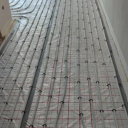 Szare rury ogrzewania podłogowego ułożone w pętle na folii izolacyjnej z czerwoną siatką w surowym wnętrzu, widok z g&oacute;ry.