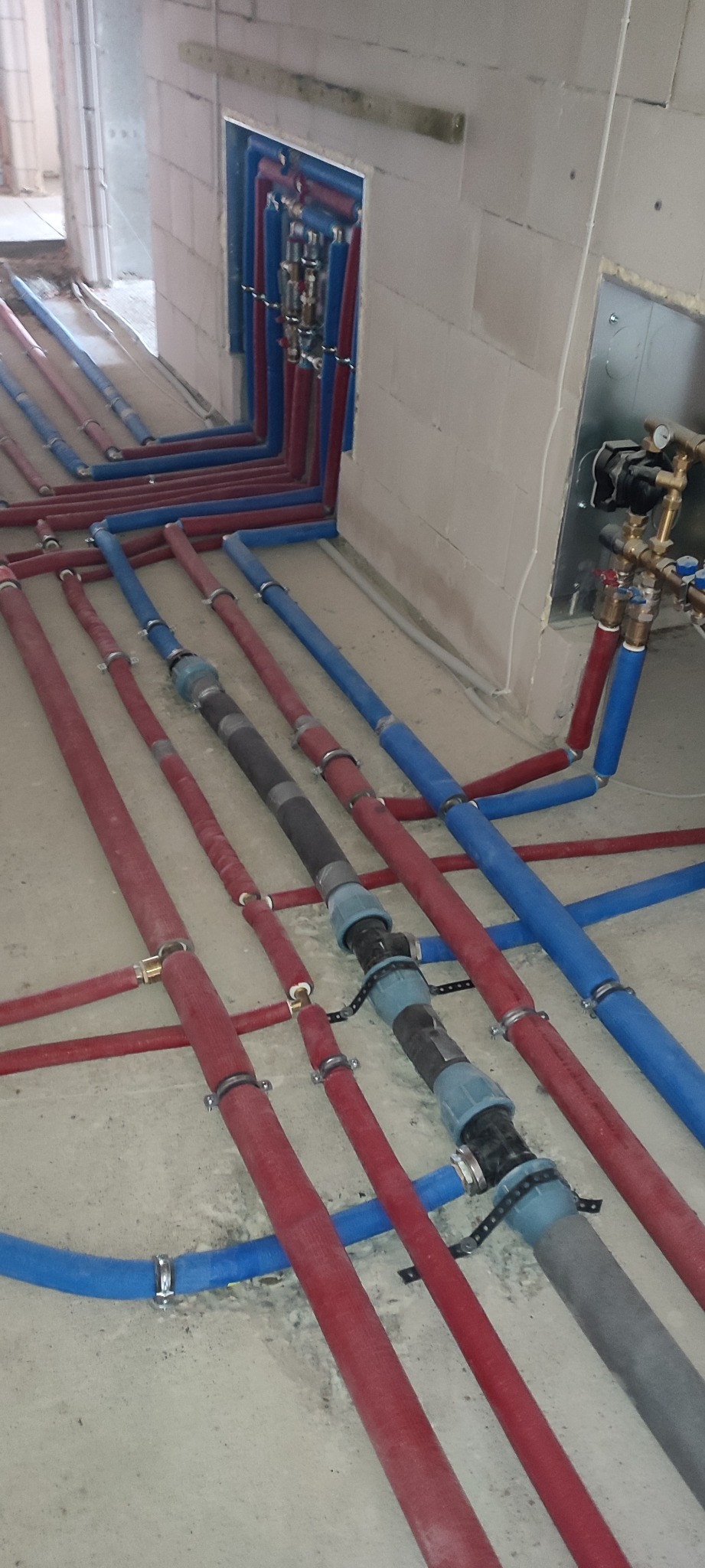 Rozprowadzenie rur instalacji wodnej i grzewczej w surowym stanie podłoża, widoczne czerwone rury z izolacją oraz niebieskie rury, złączki i kolektory.