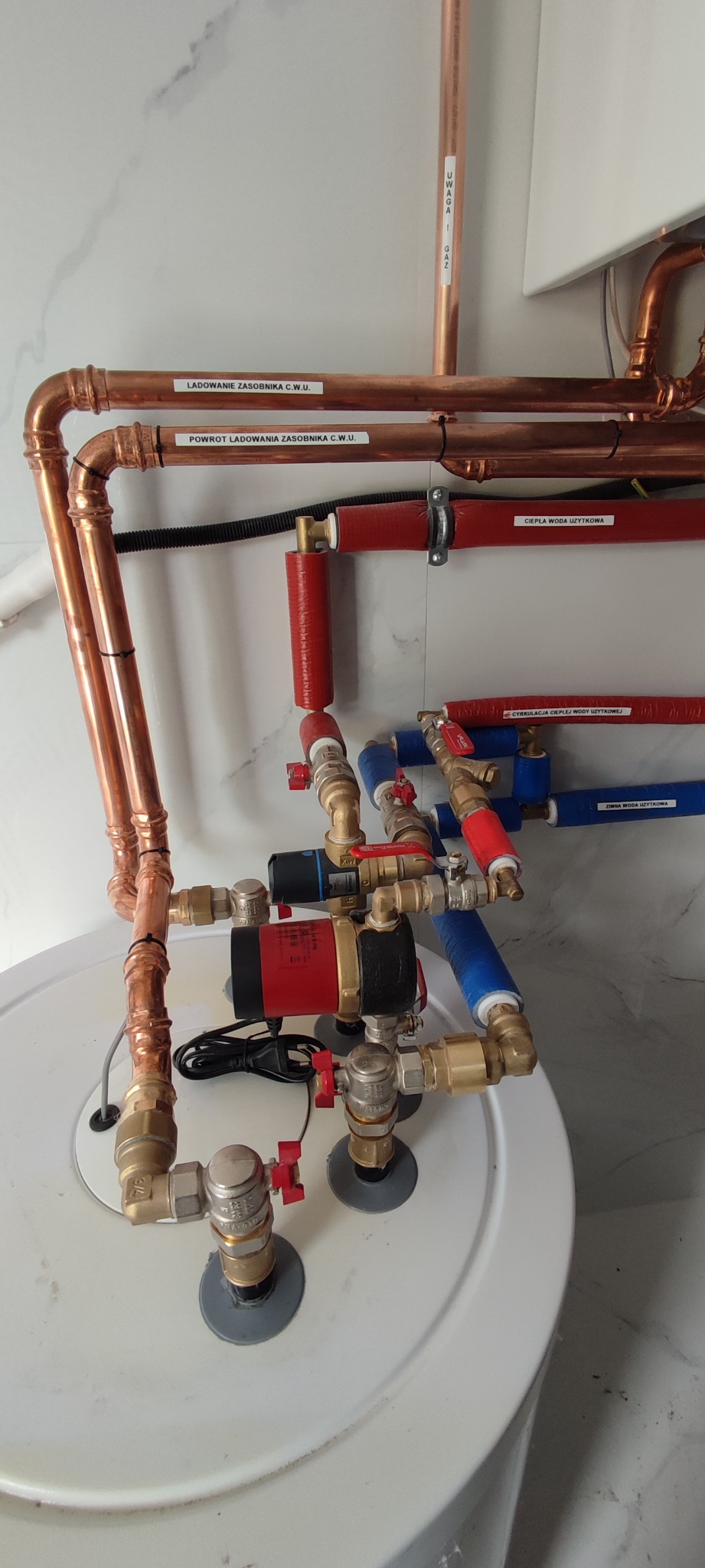 Złożona instalacja wodna z miedzianymi rurami, pompami obiegowymi i zaworami, podłączona do zasobnika ciepłej wody użytkowej, z widocznymi etykietami opisującymi funkcje poszczególnych rur...
