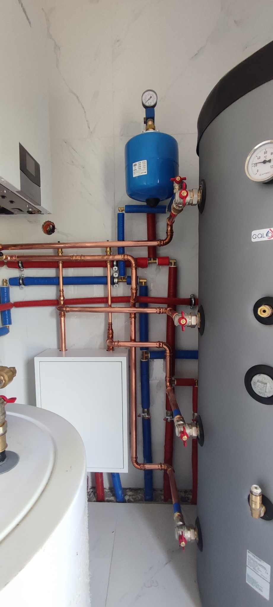 Złożona instalacja wodna z miedzianymi rurami, niebieskimi elementami i dużym szarym zbiornikiem z manometrem na tle białych płytek imitujących marmur.