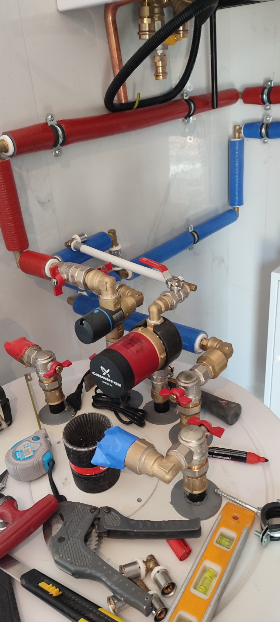 Instalacja CO z pompą obiegową Grundfos Comfort PM, rurami w izolacji czerwonej i niebieskiej, zaworami kulowymi z czerwonymi rączkami, oraz narzędziami hydraulicznymi na białym tle.