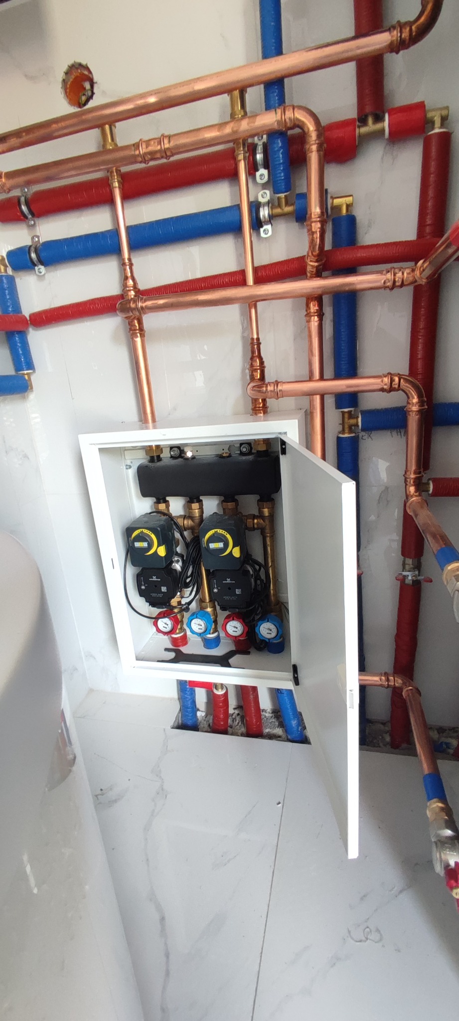 Rozdzielacz ogrzewania podłogowego w szafce z otwartymi drzwiczkami, widoczne pompy obiegowe, rury miedziane i plastikowe w kolorach czerwonym i niebieskim na tle białych płytek imitujących marmur.