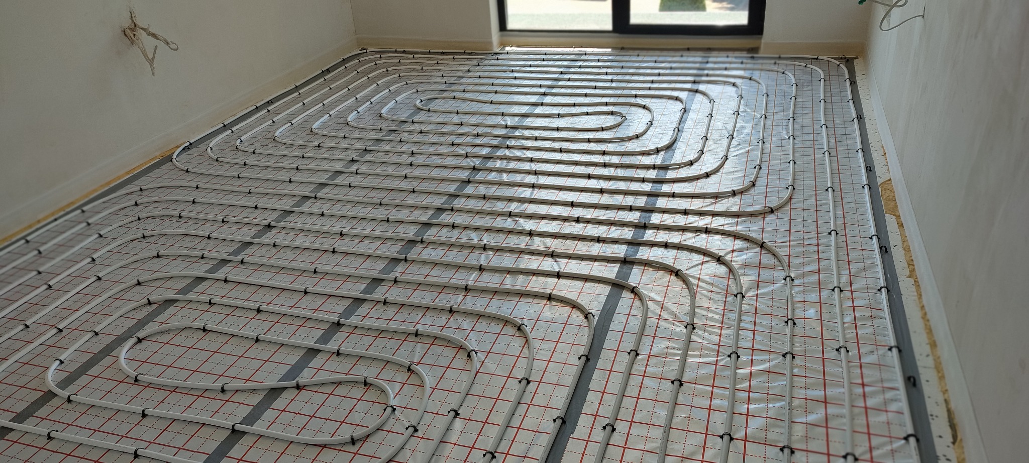 Rozłożone rury ogrzewania podłogowego na folii z siatką, widoczne czarne uchwyty mocujące, surowy stan wykończenia ścian i brak wylewki.