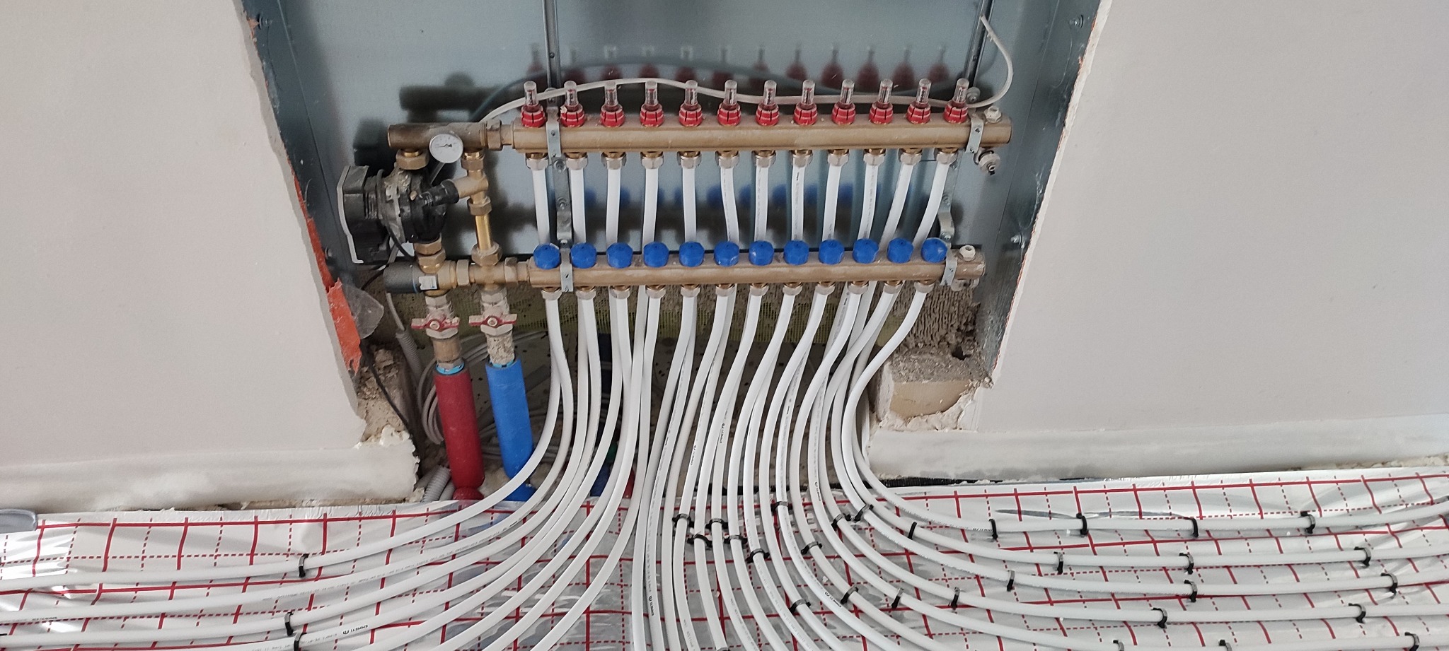 Rozdzielacz ogrzewania podłogowego z licznymi białymi rurkami, zamontowany w metalowej obudowie, widoczne czerwone i niebieskie rury doprowadzające, izolacja i mata z czerwonym nadrukiem w kratkę.