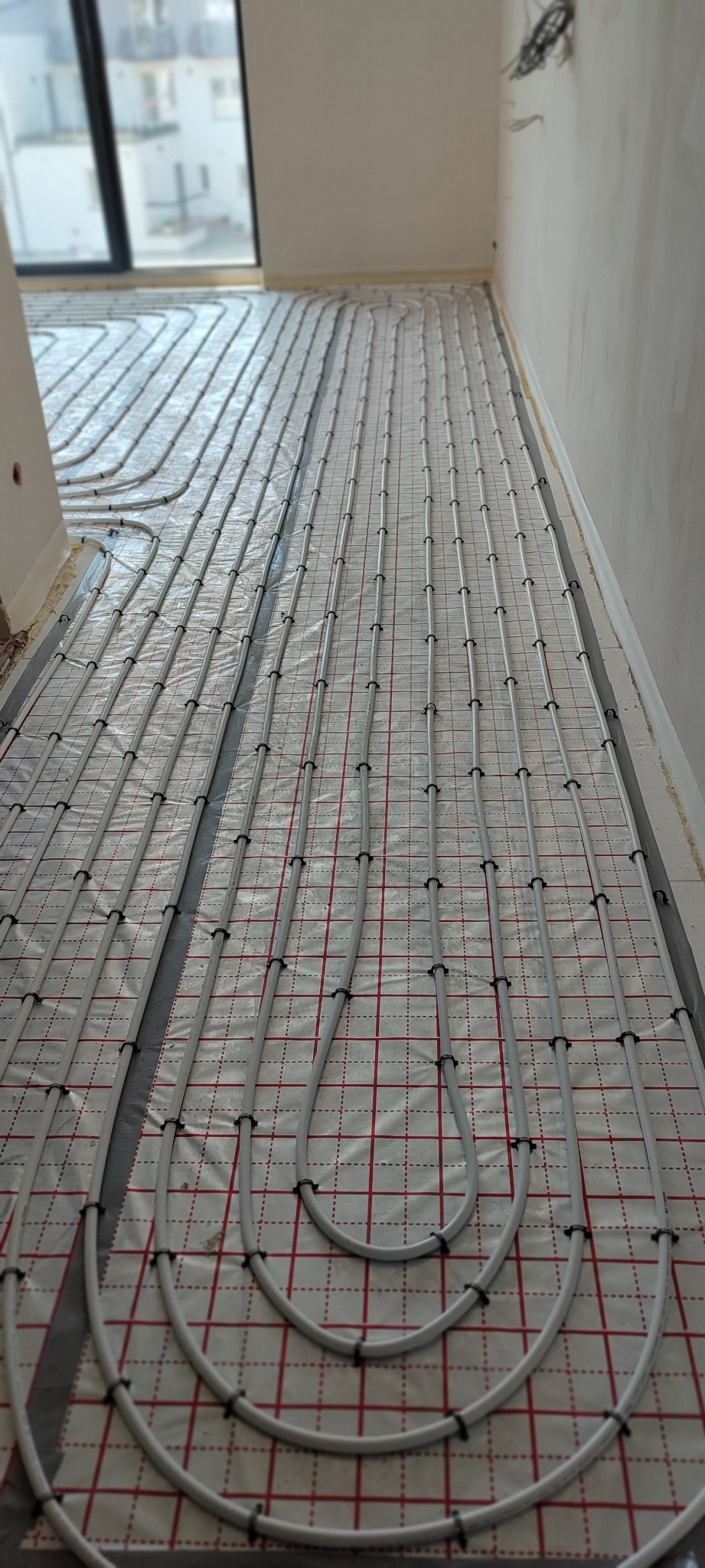 Układ rur ogrzewania podłogowego na folii z siatką w nowym budynku, widoczne mocowania i pętle rur.
