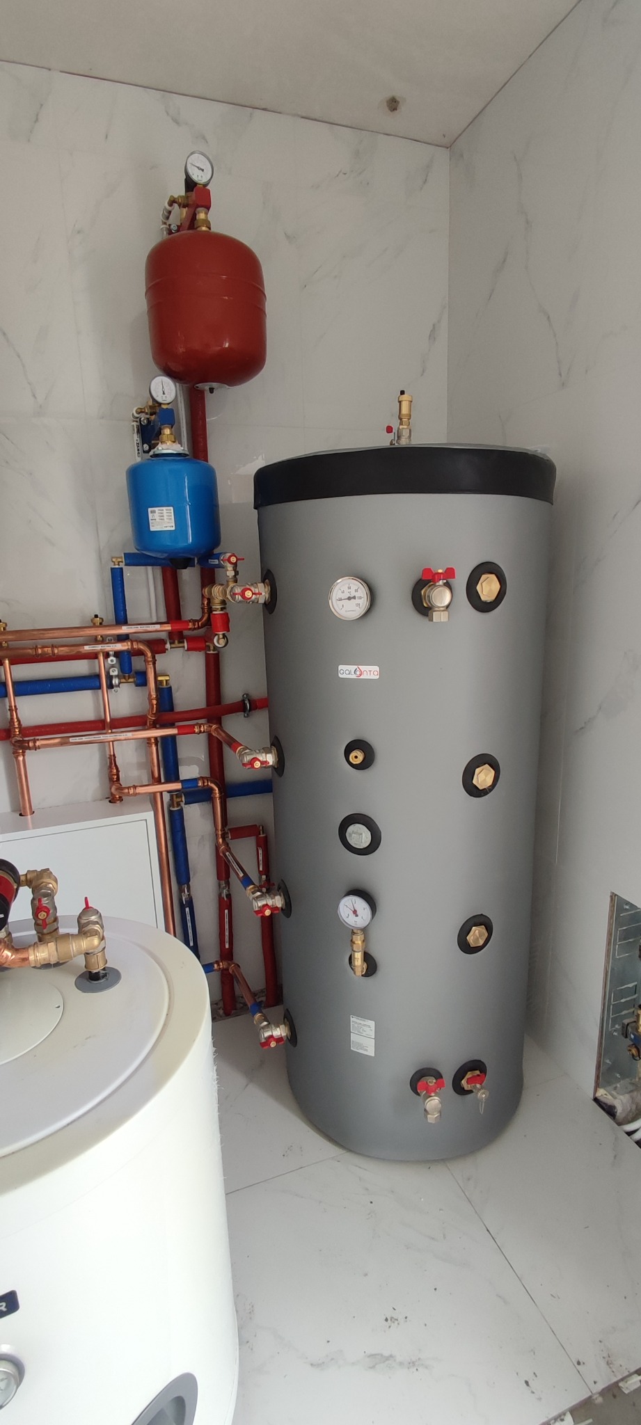 Szary zbiornik akumulacyjny ciepła z podłączonymi rurami miedzianymi i niebieskimi, czerwony zbiornik wyrównawczy na górze, całość w pomieszczeniu wykończonym białymi płytkami imitującymi marmur.