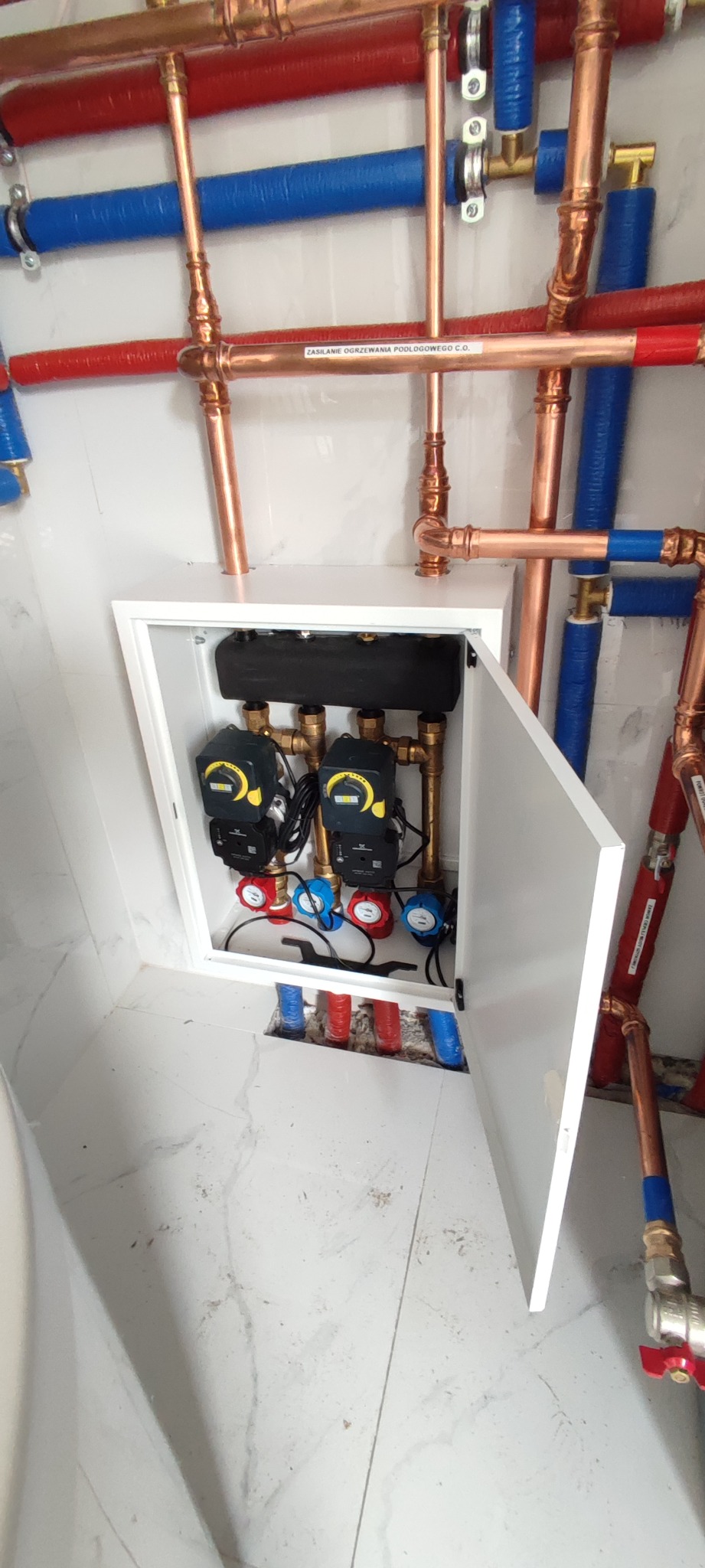 Rozdzielacz ogrzewania podłogowego w szafce, z widocznymi pompami obiegowymi i rurami miedzianymi oraz PEX w kolorach czerwonym i niebieskim, zamontowany na ścianie z marmurową okładziną.