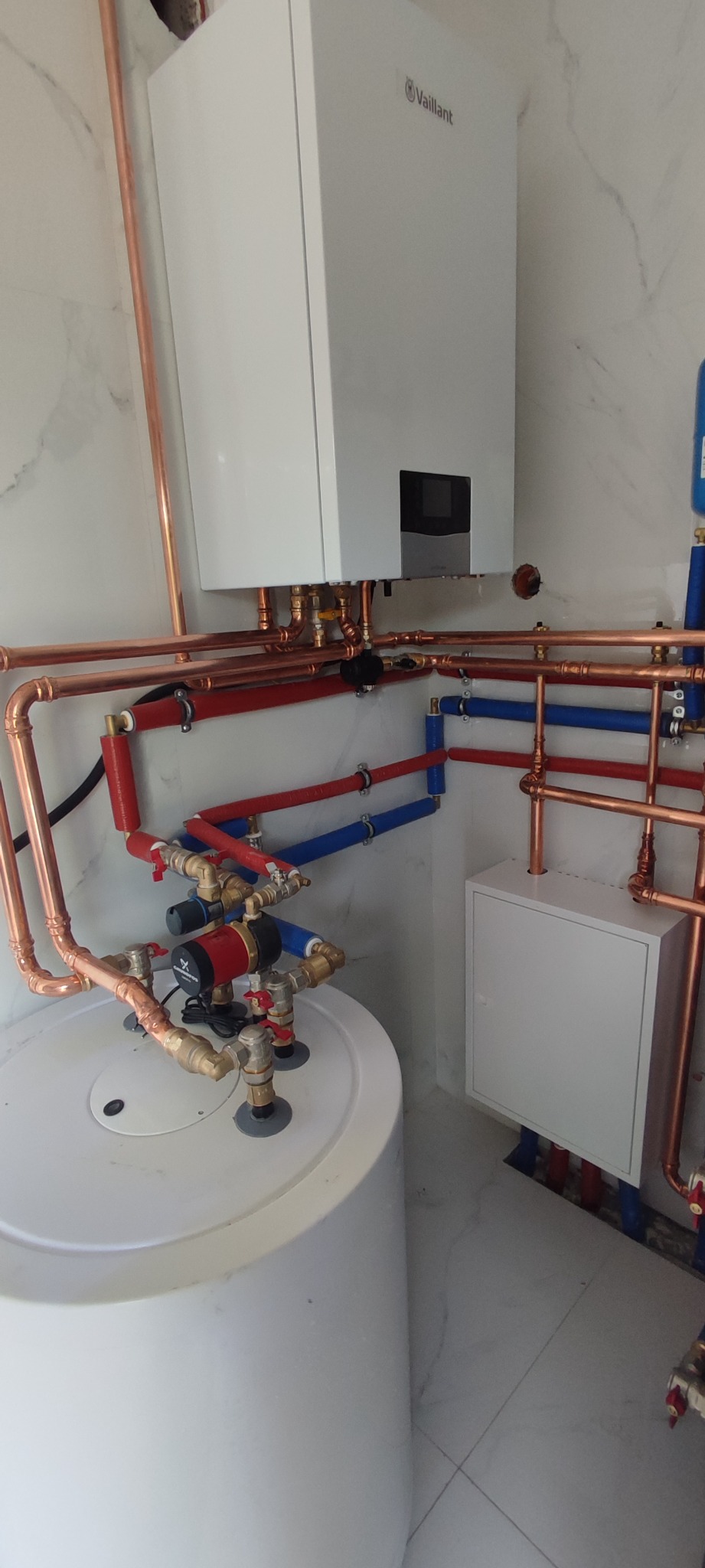 Biały piec gazowy Vaillant zamontowany na ścianie z marmurowym wzorem, z widocznymi miedzianymi rurami, pompą obiegową i zasobnikiem ciepłej wody, instalacja w pomieszczeniu technicznym.