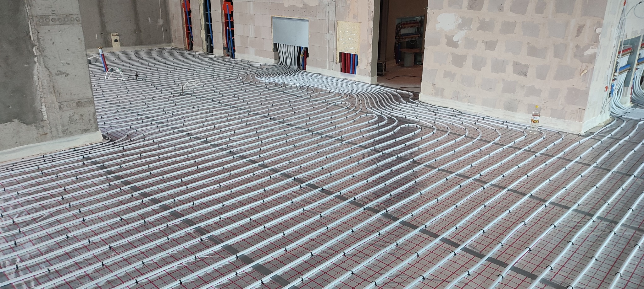 Rozłożone rury ogrzewania podłogowego na folii izolacyjnej w surowym stanie budynku, widoczne wystające kable i rury instalacji.