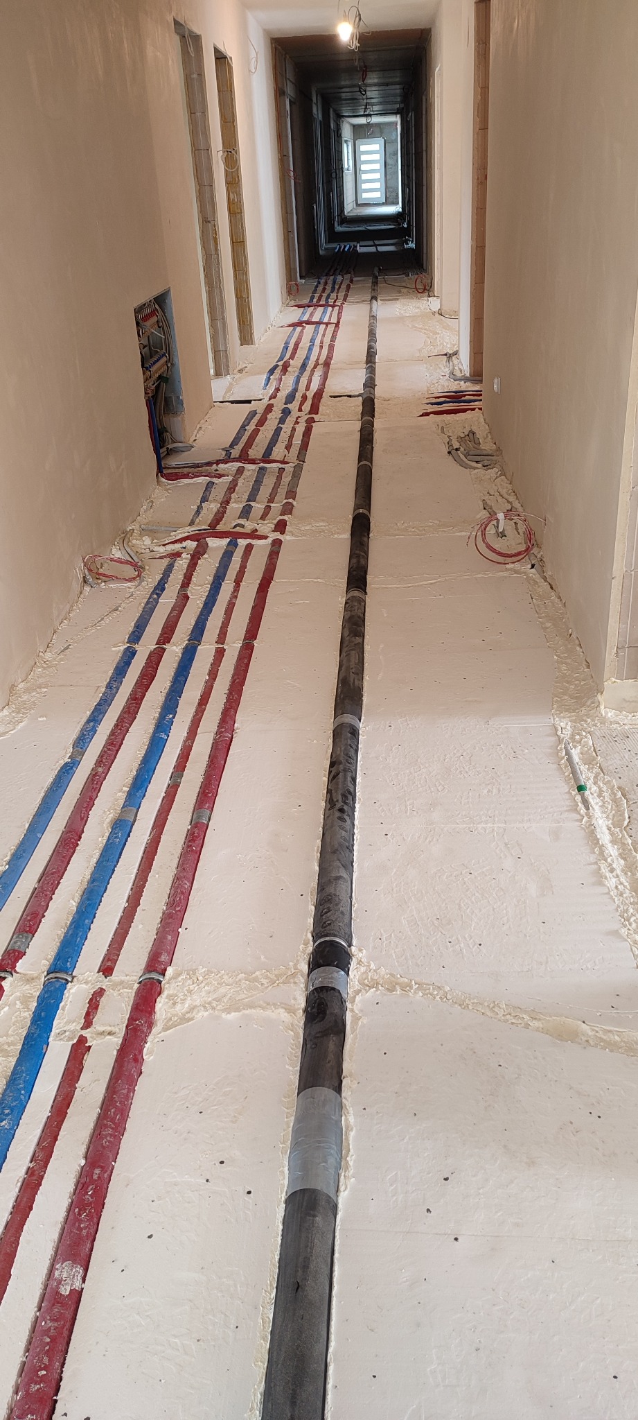 Instalacja rur grzewczych i wodociągowych w bruzdach pod wylewką, widok w perspektywie długiego korytarza w surowym stanie.