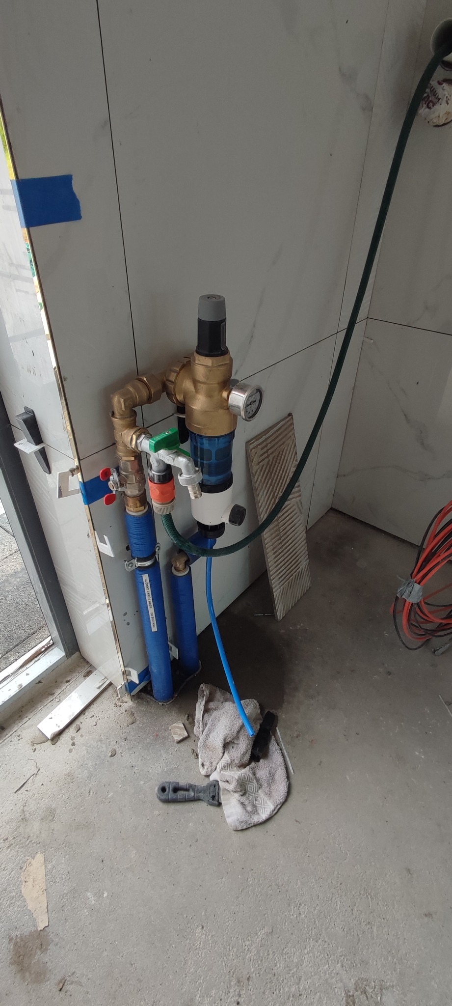 Szczegółowe ujęcie podłączenia instalacji wodnej z filtrem, zaworami i manometrem, widoczne niebieskie rury i mosiężne złączki na tle białych płytek, z elementami wykończeniowymi w trakcie montażu.