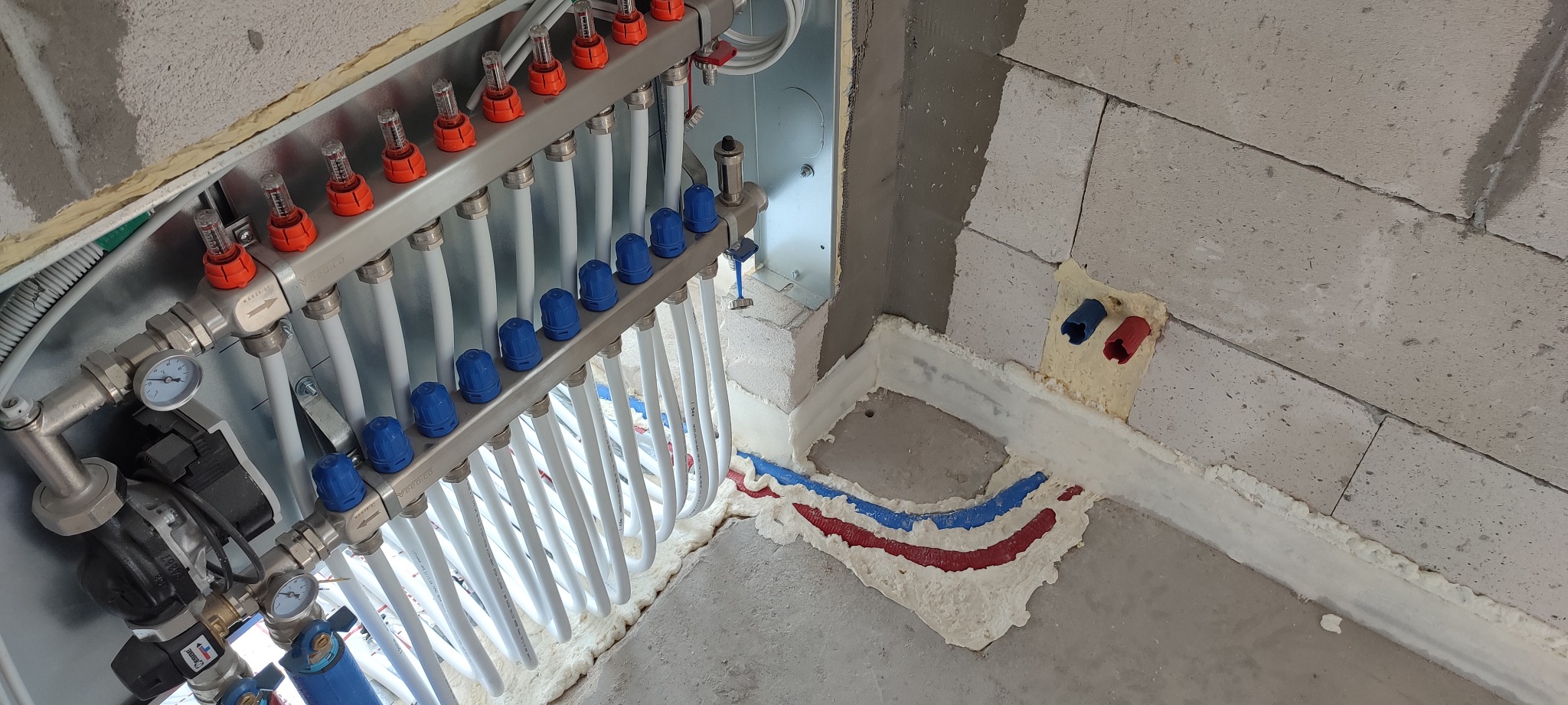 Rozdzielacz ogrzewania podłogowego z białymi rurami, zamontowany w surowym wnętrzu z betonową posadzką i ścianami z bloczków, zabezpieczony pianką montażową.