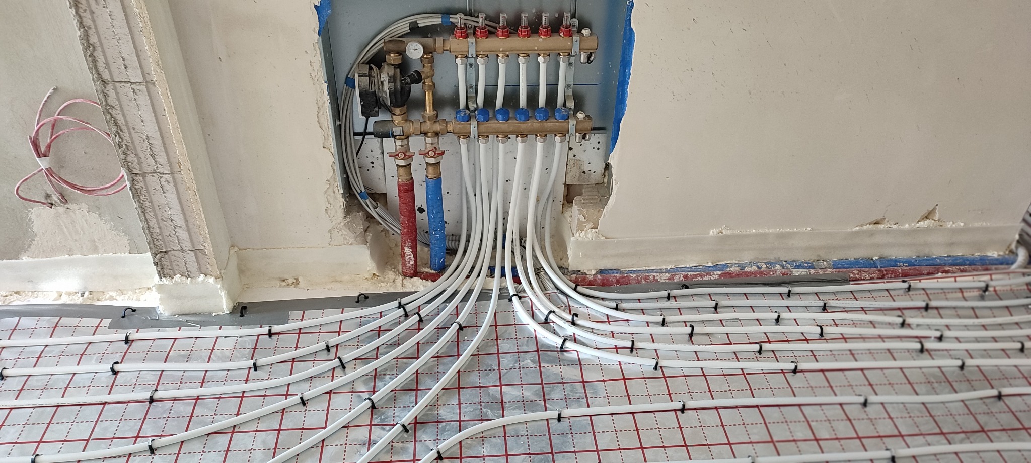Rozdzielacz ogrzewania podłogowego z białymi rurami ułożonymi na folii z czerwoną siatką, widoczne przygotowania do zalania wylewką, surowy stan ścian.
