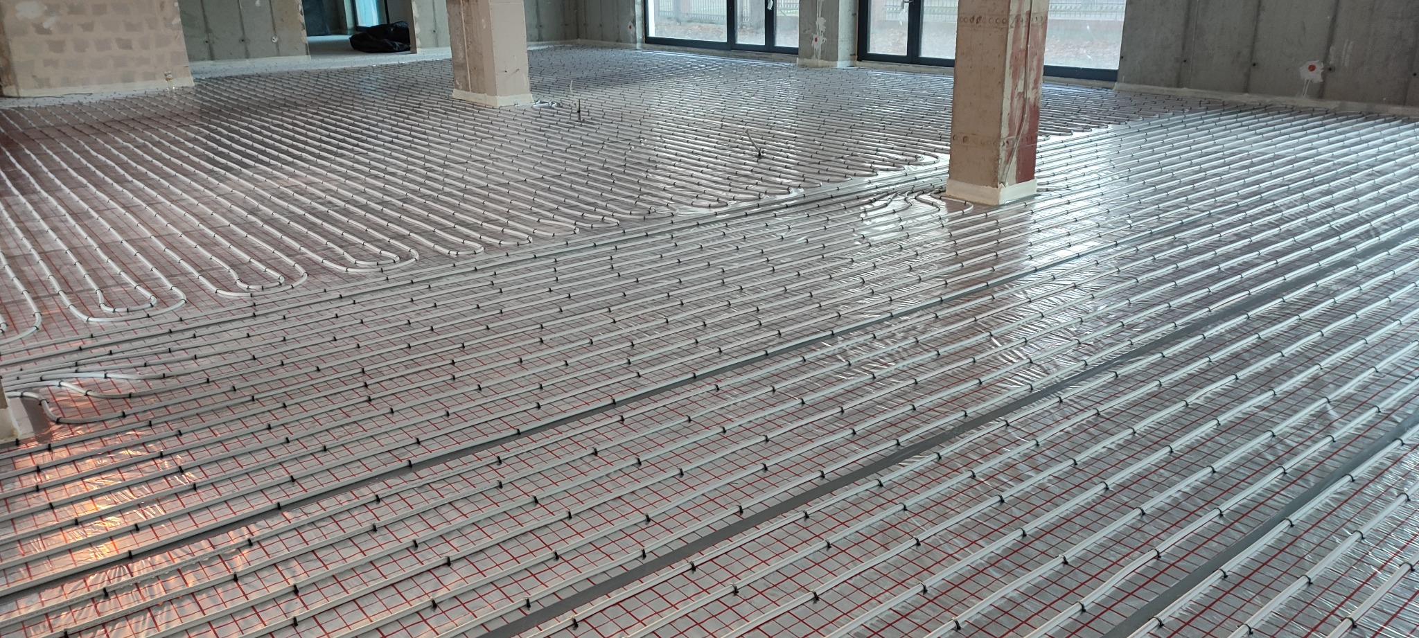 Rozległy system ogrzewania podłogowego w trakcie montażu, rury ułożone równolegle na izolacji z czerwoną siatką, widoczne słupy konstrukcyjne w surowym stanie.