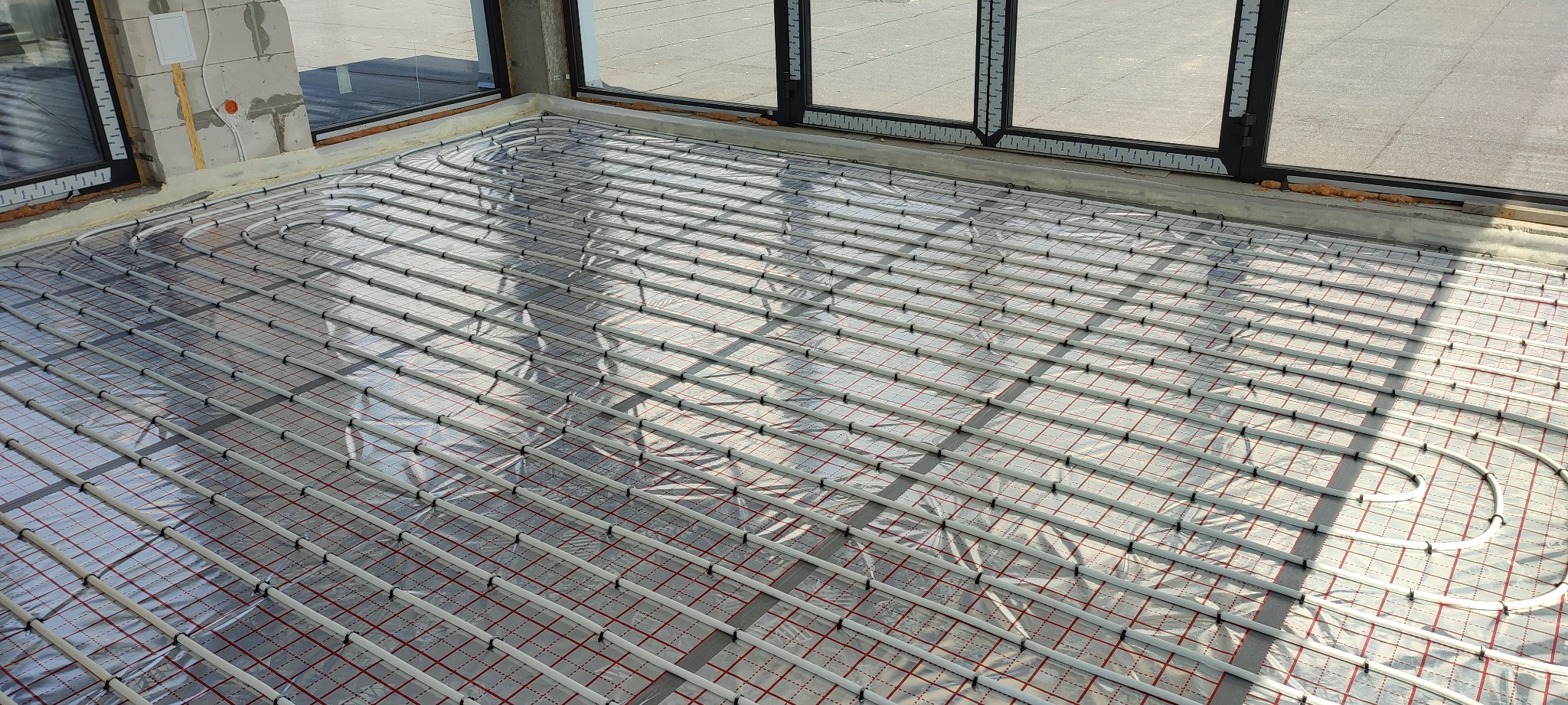 Rozłożone rury ogrzewania podłogowego na folii izolacyjnej z rastrem, widoczne okna i fragment ściany z instalacją elektryczną w surowym stanie.