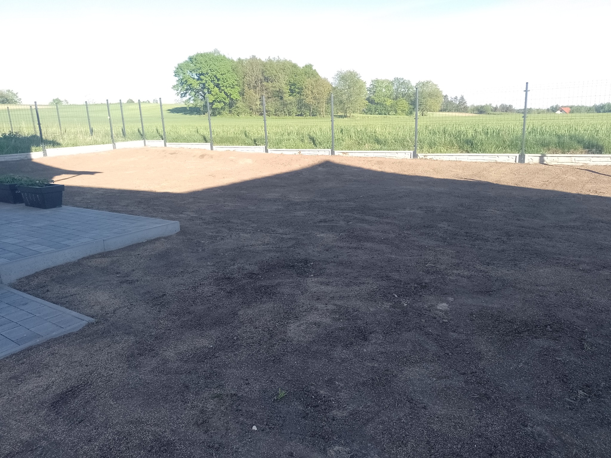 Przygotowany teren pod trawnik z obrzeżami, widok na siatkę ogrodzeniową i pole w tle, wczesna faza zakładania ogrodu.