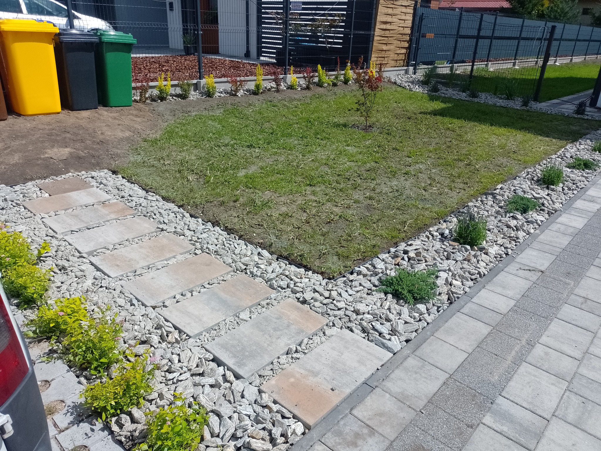 Świeżo założony trawnik z młodymi drzewkami i krzewami, obrzeża wysypane białym grysem, widoczne ścieżki z płyt chodnikowych, w tle pojemniki na śmieci.