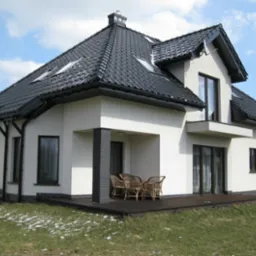 Nowoczesny dom jednorodzinny z jasną elewacją, ciemną dachówką i drewnianym tarasem, widok z ogrodu w słoneczny dzień.