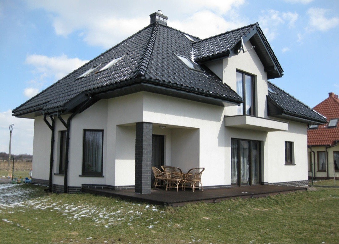 Nowoczesny dom jednorodzinny z jasną elewacją, ciemną dachówką i drewnianym tarasem, widok z ogrodu w słoneczny dzień.