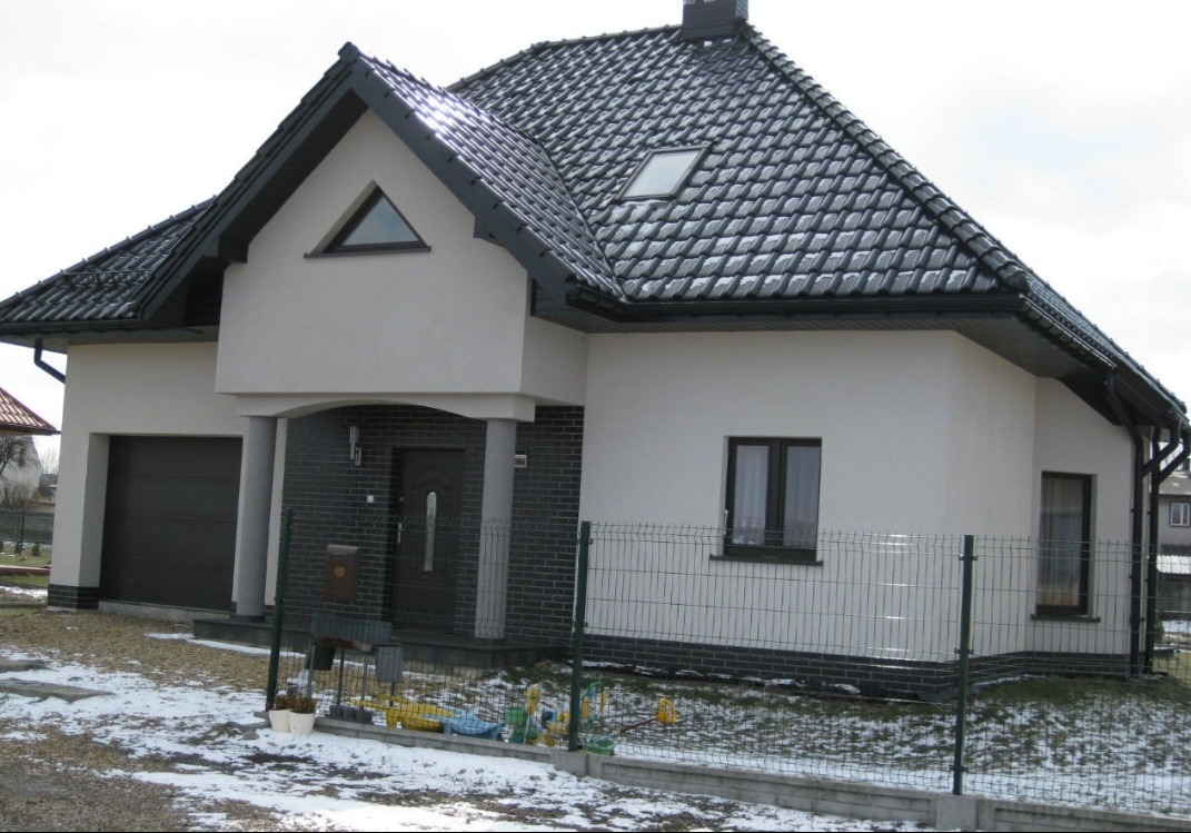 Nowoczesny dom jednorodzinny z białą elewacją, ciemnoszarą dachówką i elementami z ciemnej cegły, ogrodzony siatką na tle resztek śniegu.