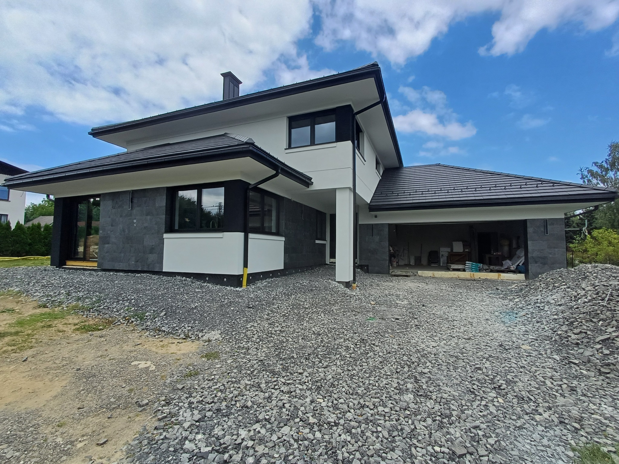 Nowoczesny dom jednorodzinny z grafitową elewacją częściowo wykończoną kamieniem, czarną stolarką okienną i dachem, widoczne przygotowania do zagospodarowania terenu wokół budynku, wysypany żwir.