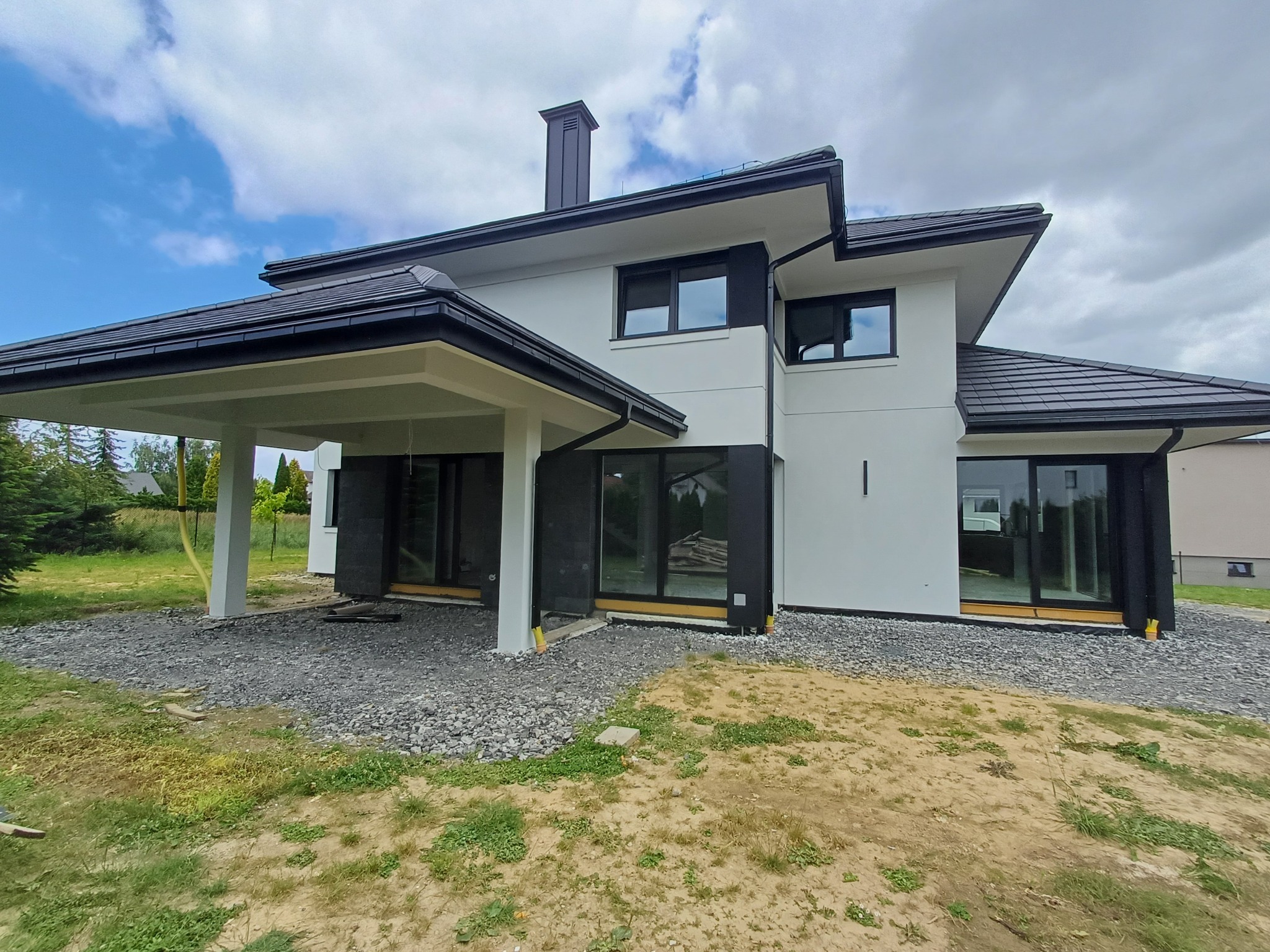 Nowoczesny dom z białymi ścianami i czarnymi obramowaniami okien, widoczne szare kamienie wokół budynku, błękitne niebo z chmurami w tle.