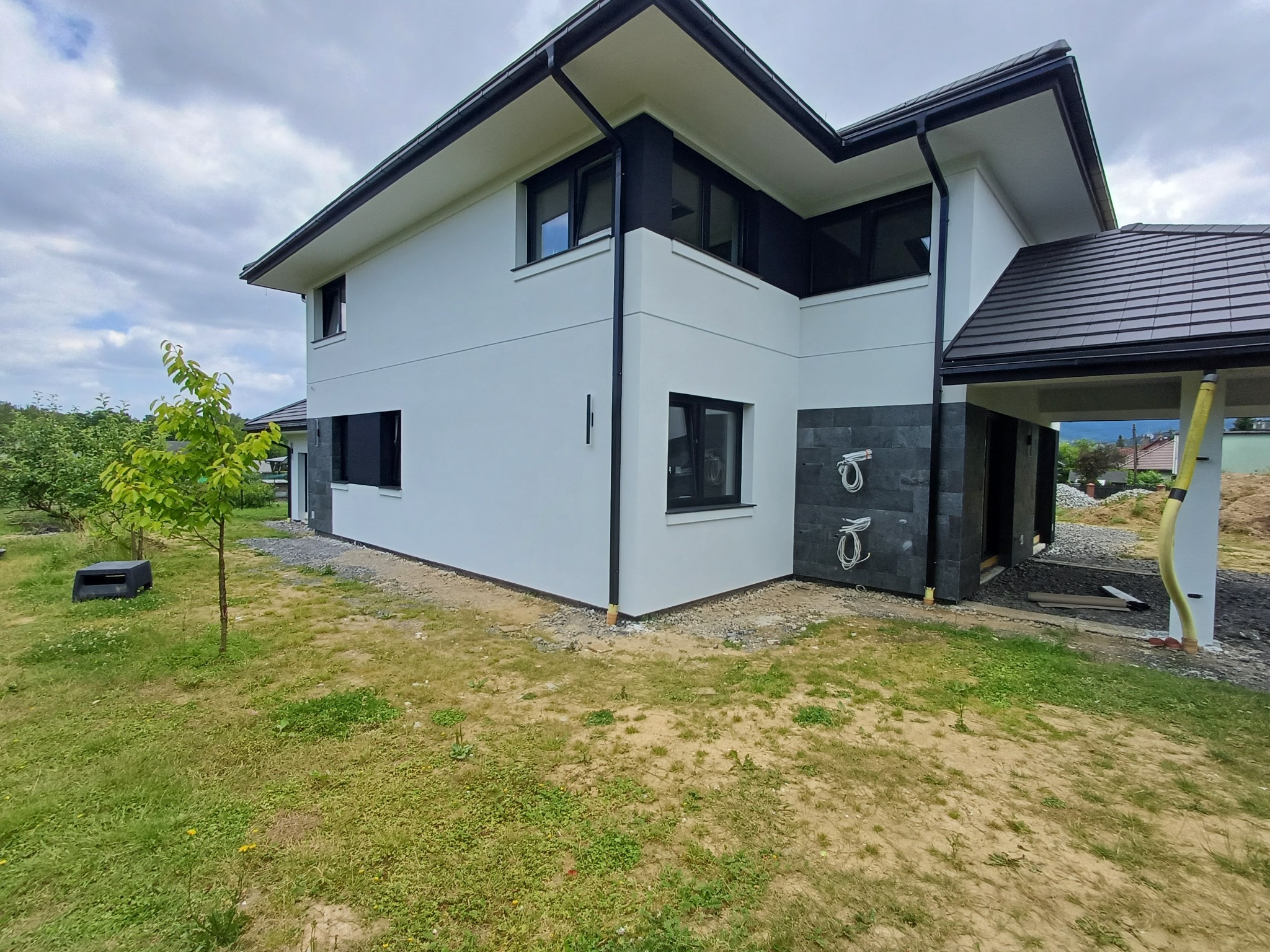 Dwukondygnacyjny dom z białą elewacją i czarnymi elementami architektonicznymi, widok z zewnątrz na trawnik i fragment ogrodu.