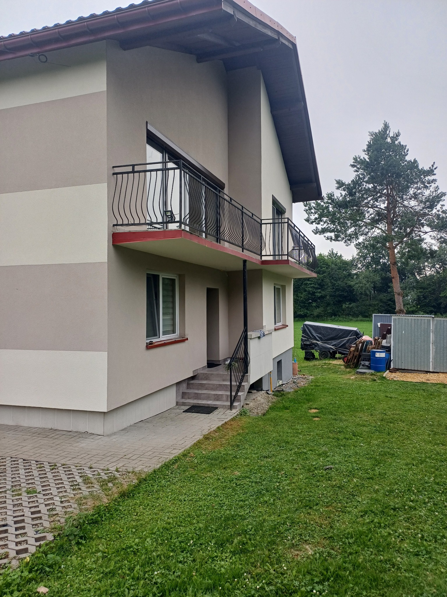 Dwukondygnacyjny dom z balkonami, elewacja w odcieniach beżu i szarości, widok z trawnika, w tle wysokie drzewo iglaste.