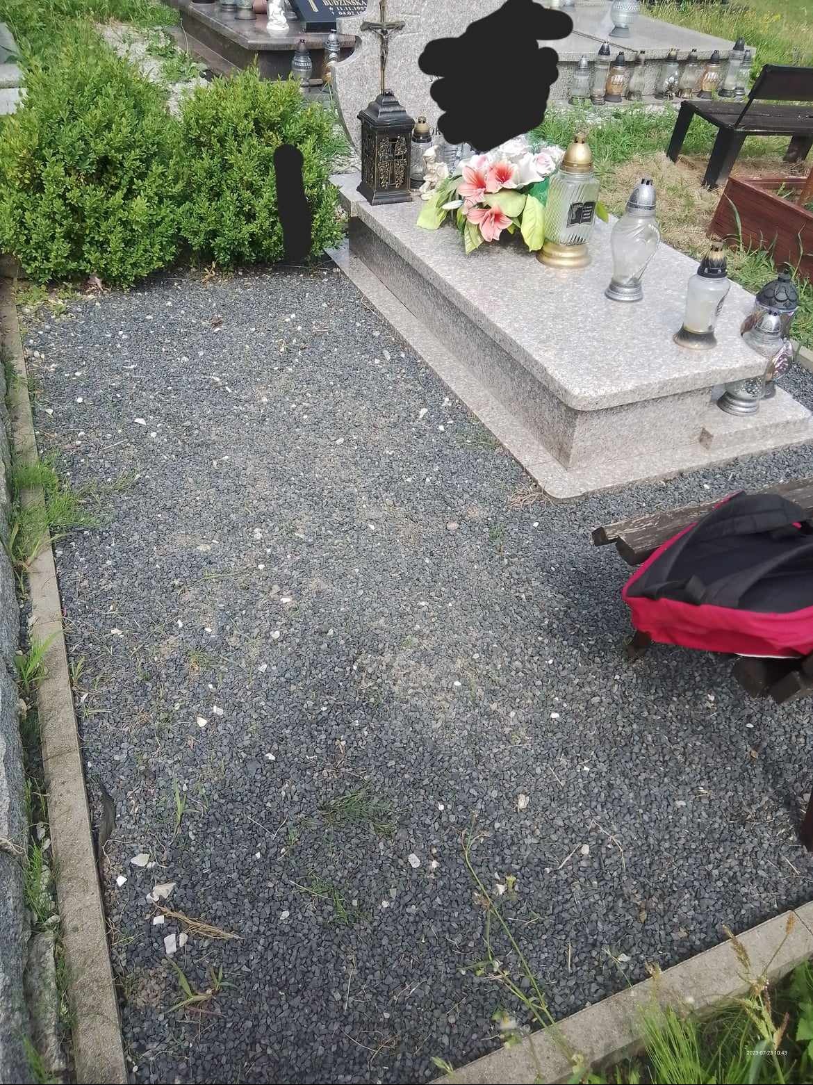 Nagrobek z kwiatami i lampionami, otoczony szarym żwirem i niskim żywopłotem, z fragmentem ławki i plecaka widocznym w prawym dolnym rogu.