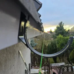 Montaż rynny dachowej z widoczną poziomicą opartą o ścianę budynku, w tle zieleń i zachodzące słońce.