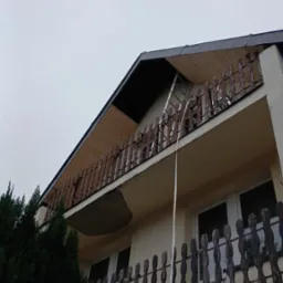 Poddasze domu z widocznym ociepleniem od zewnątrz, drabina oparta o balkon z drewnianą balustradą, fragment elewacji z widocznym ubytkiem tynku.