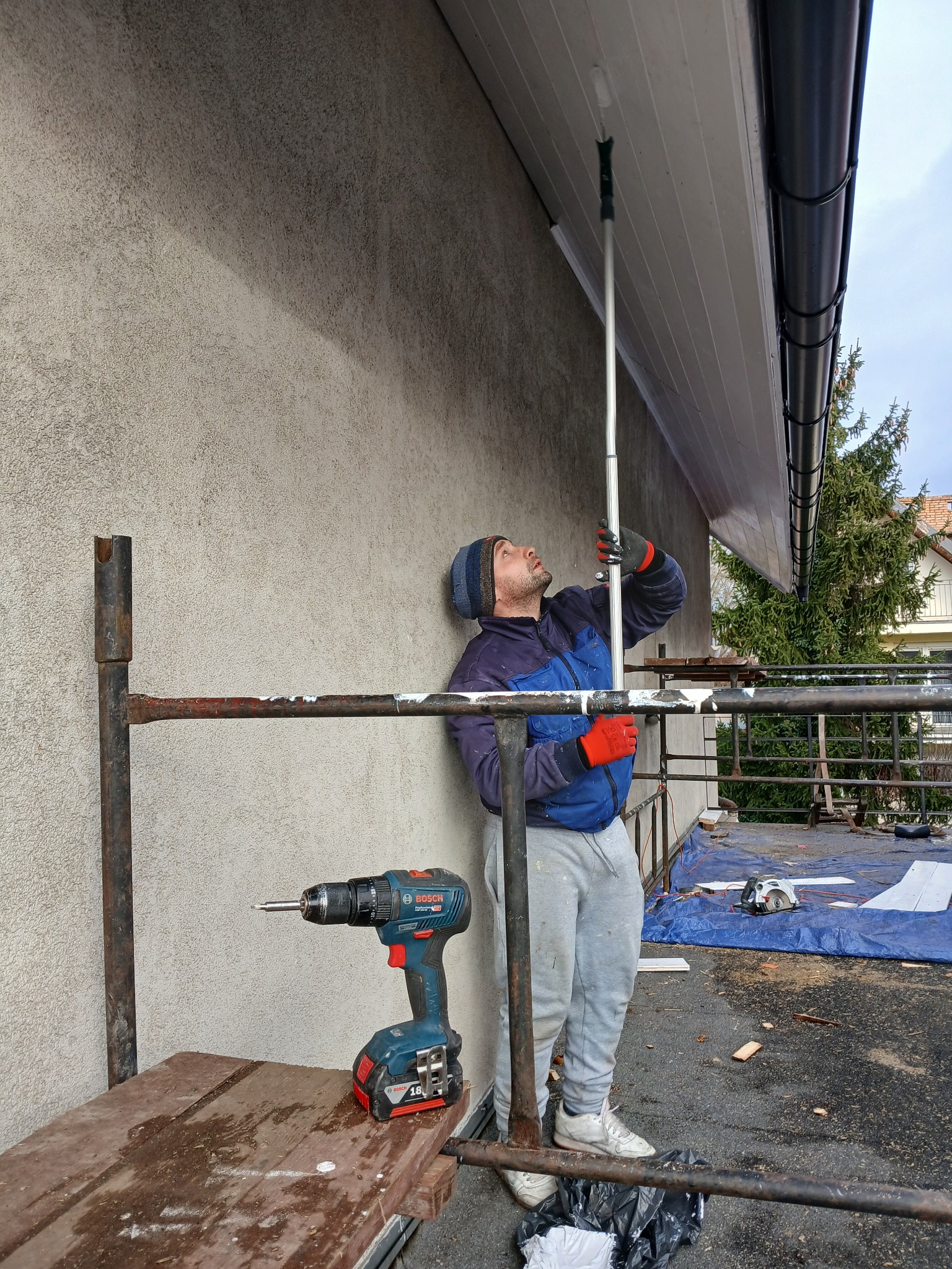 Pracownik na rusztowaniu, w czerwonych rękawicach, montuje podbitkę dachową za pomocą przedłużonego narzędzia; w tle ocieplona ściana budynku i iglaste drzewo.