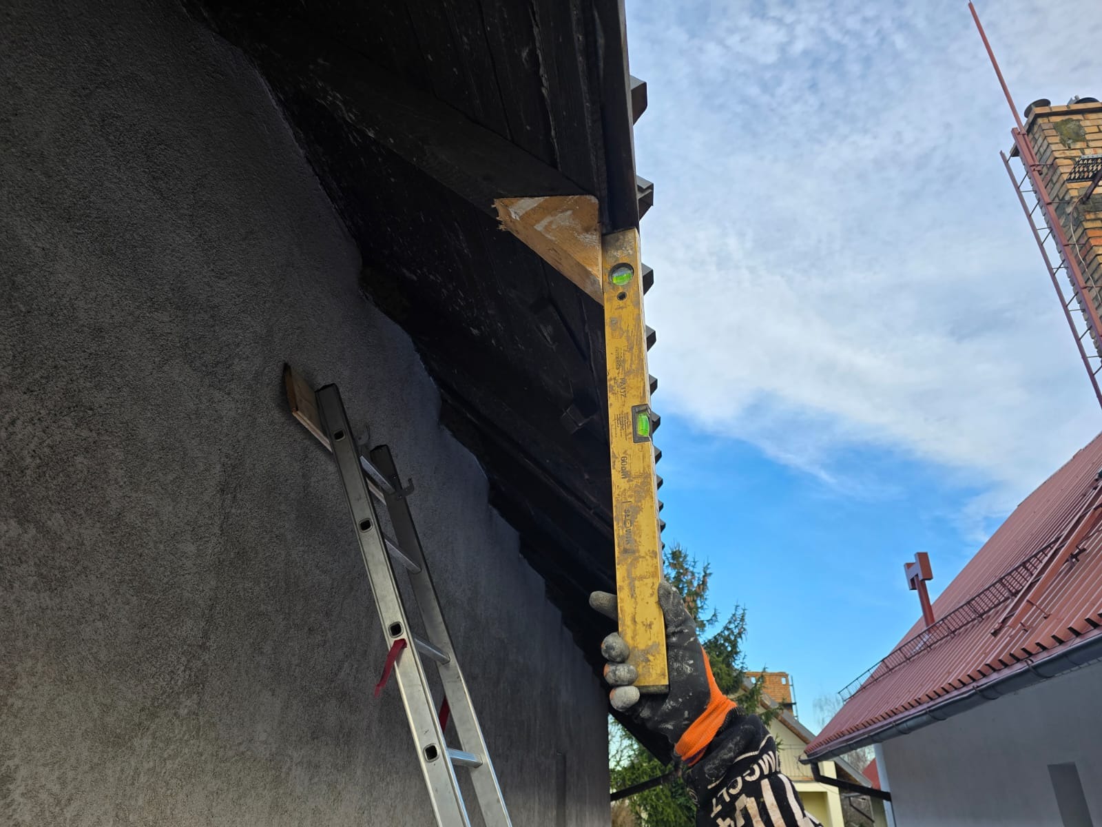 Praca z poziomicą przy ocieplaniu poddasza w Lwówku Śląskim, widoczna drabina oparta o ścianę budynku z szarą elewacją, niebo w tle.