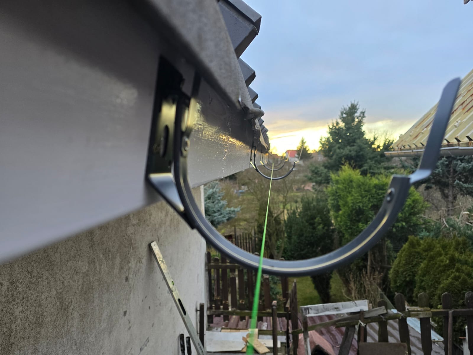 Montaż rynny dachowej z widoczną poziomicą opartą o ścianę budynku, w tle zieleń i zachodzące słońce.