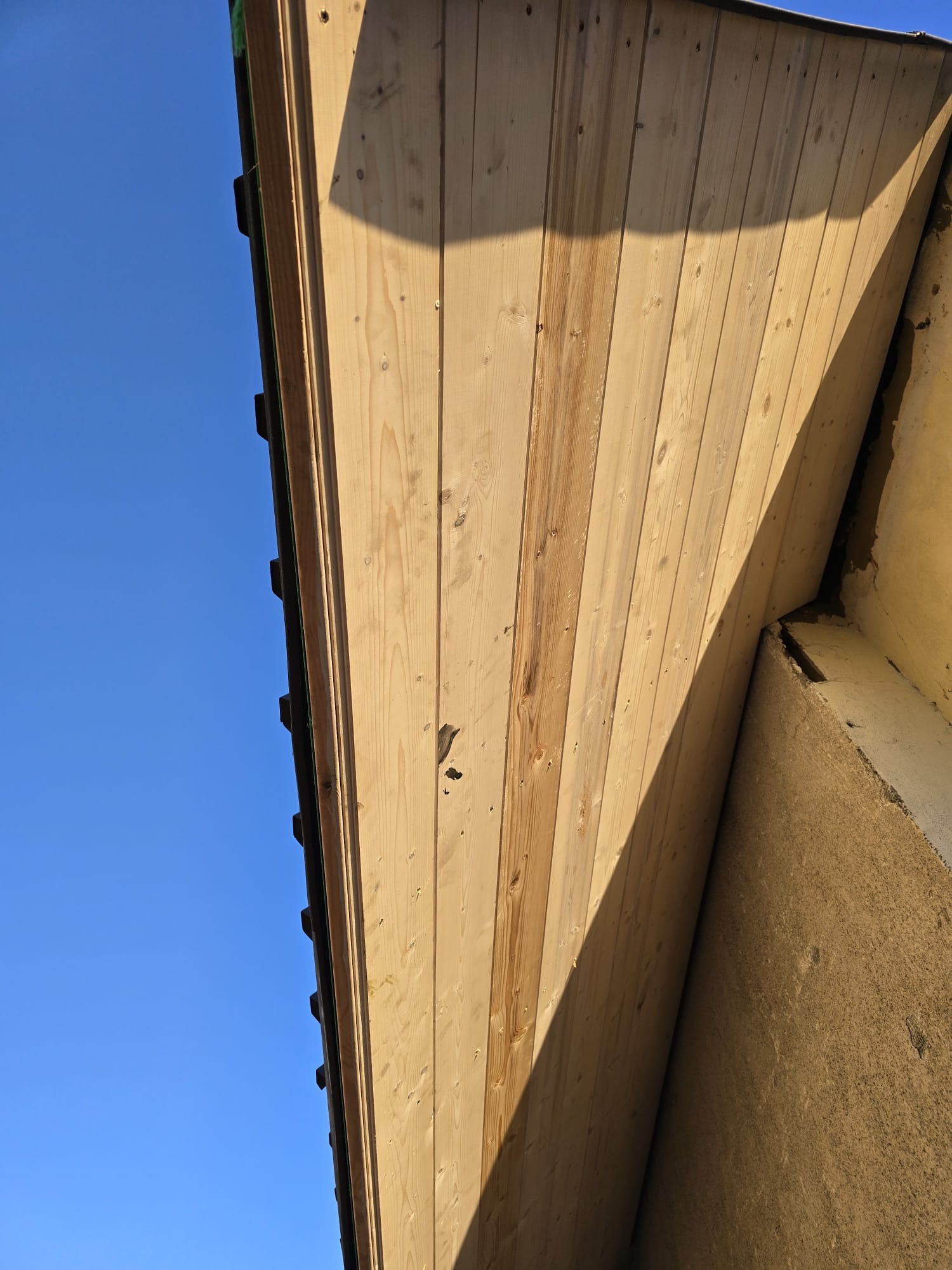 Wykończone drewnianą boazerią poddasze, widok od dołu na tle błękitnego nieba, widoczne mocowania i fragment ściany budynku.