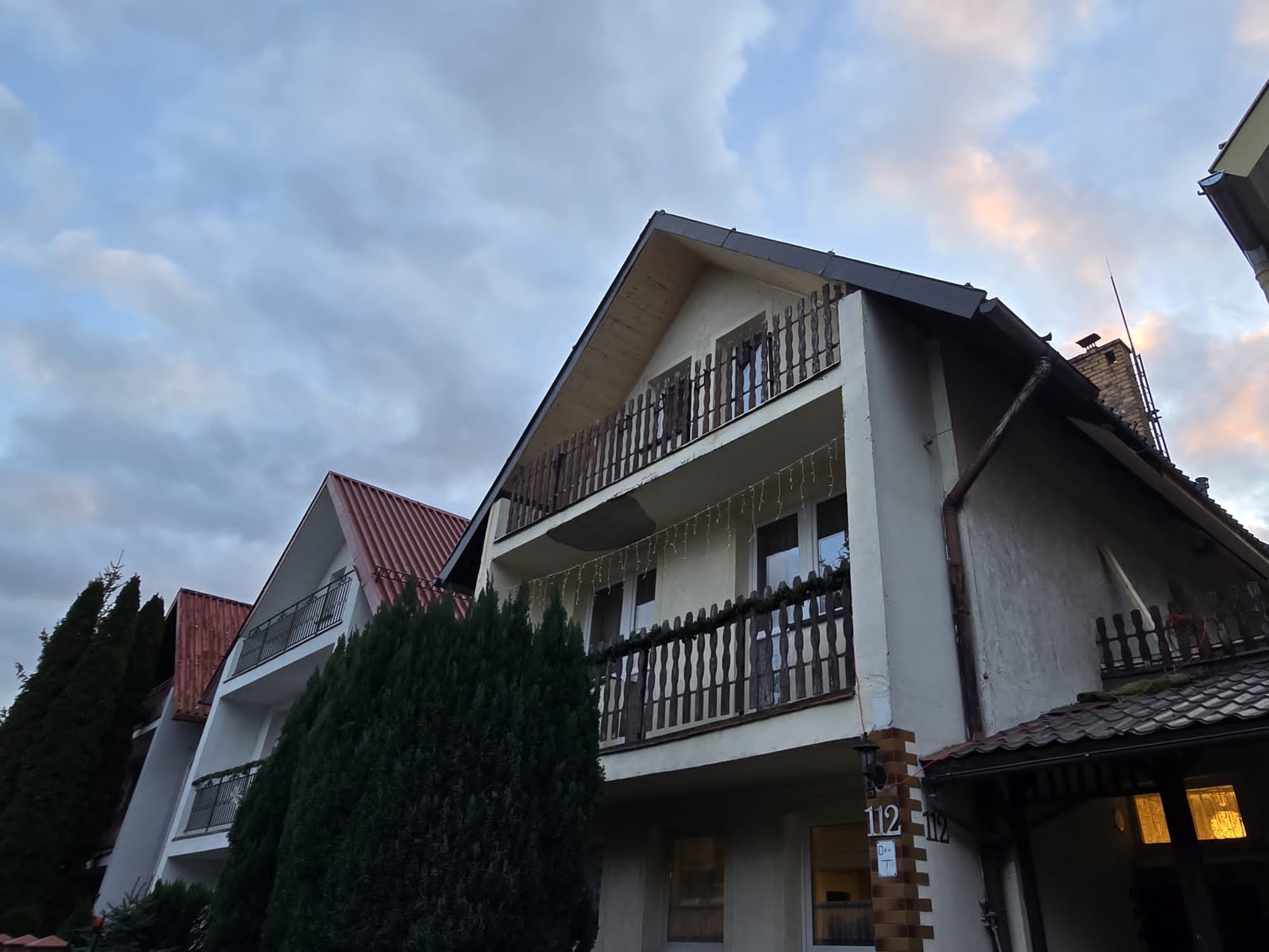 Widok na trzy budynki z balkonami, z których jeden ma drewniane balustrady i świąteczne lampki, a elewacja wymaga odświeżenia.