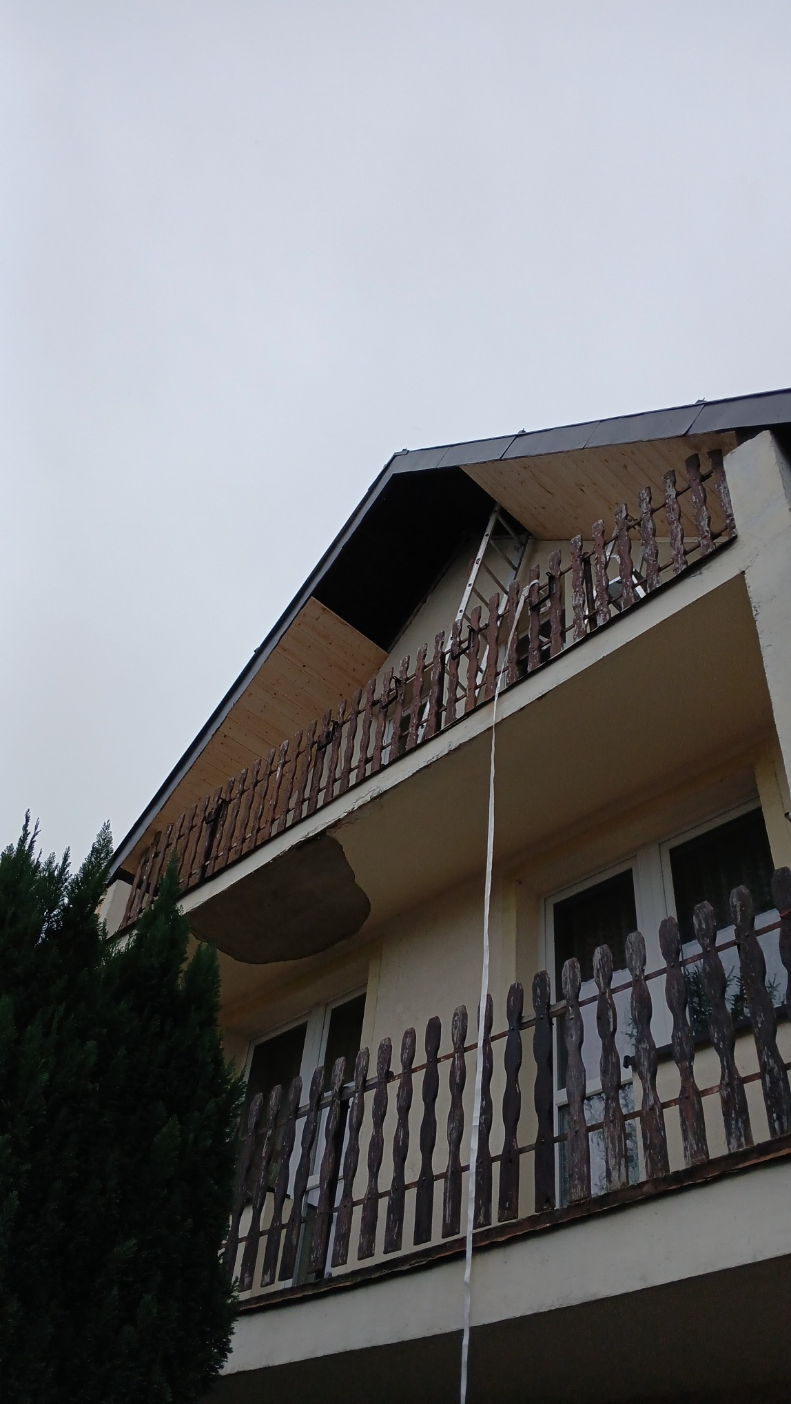 Poddasze domu z widocznym ociepleniem od zewnątrz, drabina oparta o balkon z drewnianą balustradą, fragment elewacji z widocznym ubytkiem tynku.