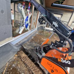 GLASSLIFT SPÓŁKA Z OGRANICZONĄ ODPOWIEDZIALNOŚCIĄ - Pomarańczowy mini-dźwig gąsienicowy BG Lift M060 z przyssawkami, montujący dużą szybę w nowym budynku w trakcie budowy.