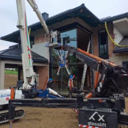 Montaż okna w domu jednorodzinnym przy użyciu podnośnika koszowego z przyssawkami. Dwóch pracowników w kaskach nadzoruje proces.