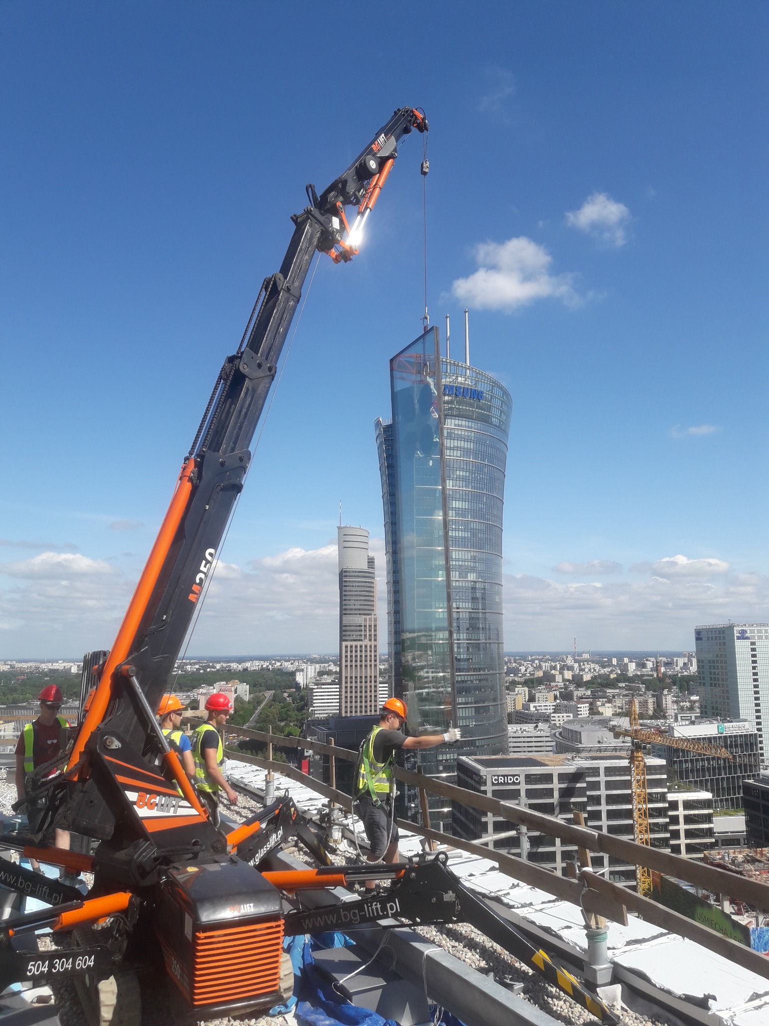 Montaż szklanej fasady wieżowca w Warszawie za pomocą dźwigu teleskopowego M250, widoczni pracownicy w kaskach i kamizelkach odblaskowych, panorama miasta w tle.
