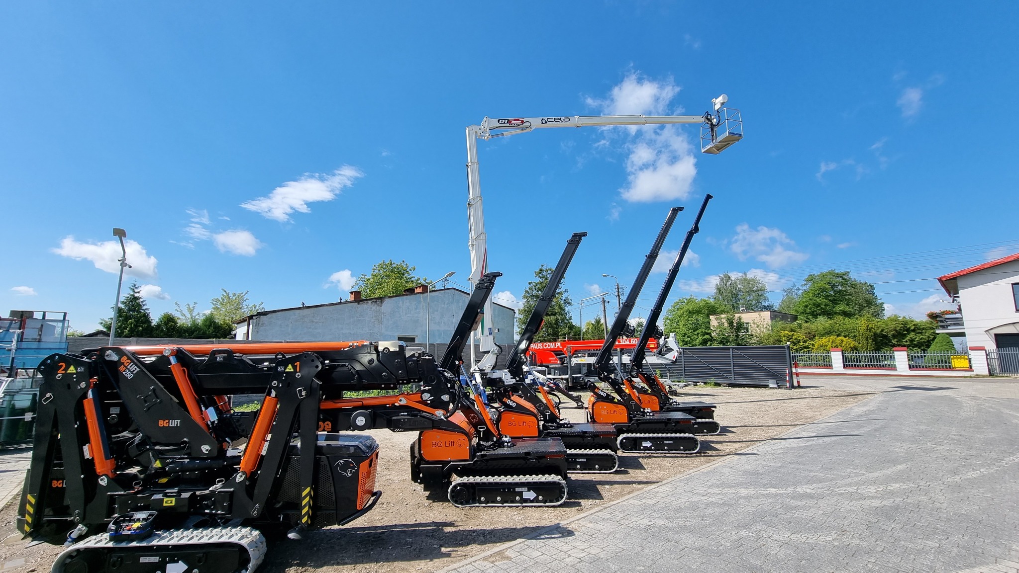 Rząd mini-dźwigów gąsienicowych w kolorach czarnym i pomarańczowym, zaparkowanych na placu pod błękitnym niebem z lekkimi chmurami, jeden z nich ma wysunięte ramię z koszem na końcu.