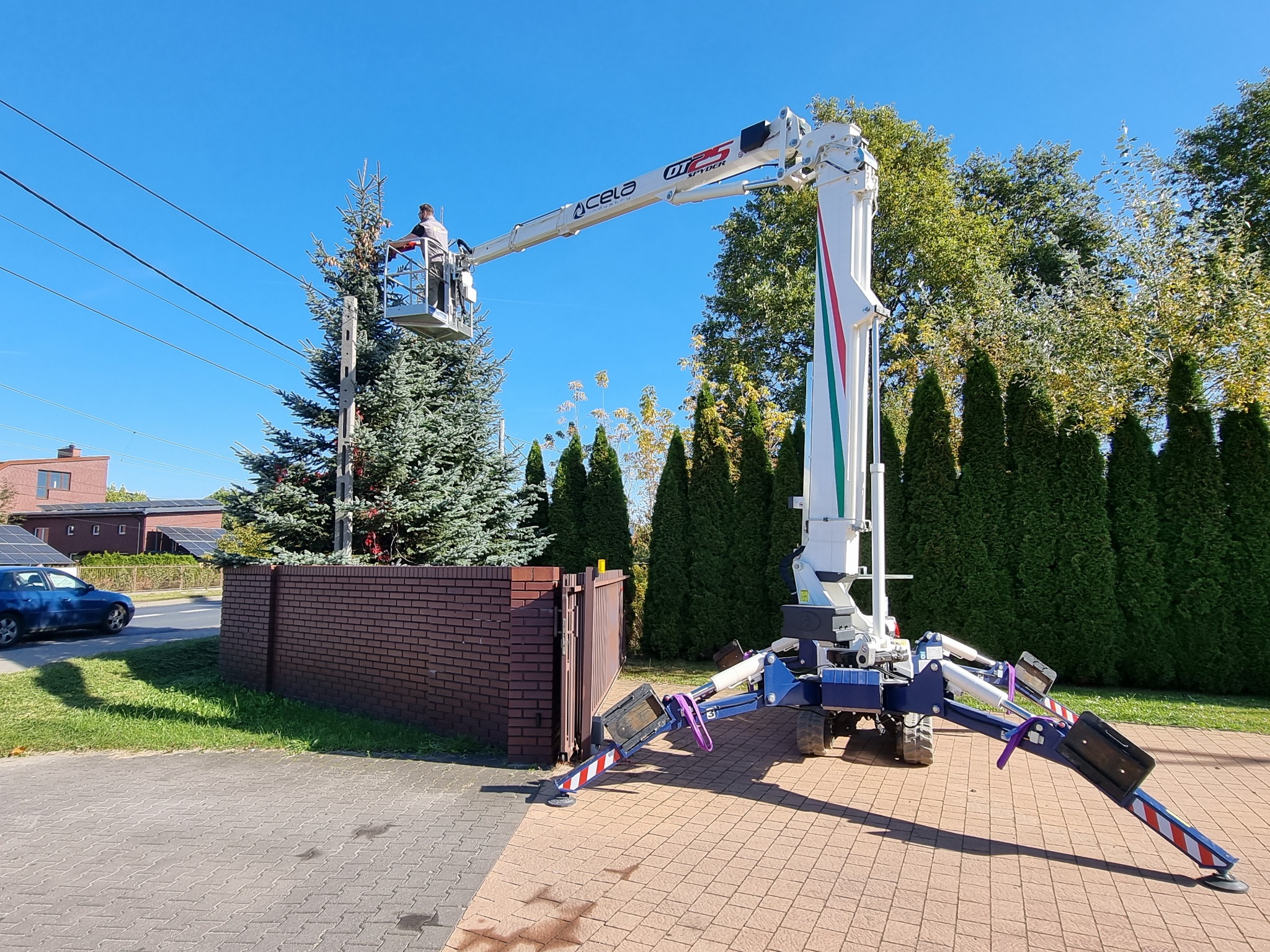 Pracownik w koszu podnośnika teleskopowego, przycina gałęzie wysokiego świerku w pobliżu linii energetycznych, na tle błękitnego nieba i zabudowań.