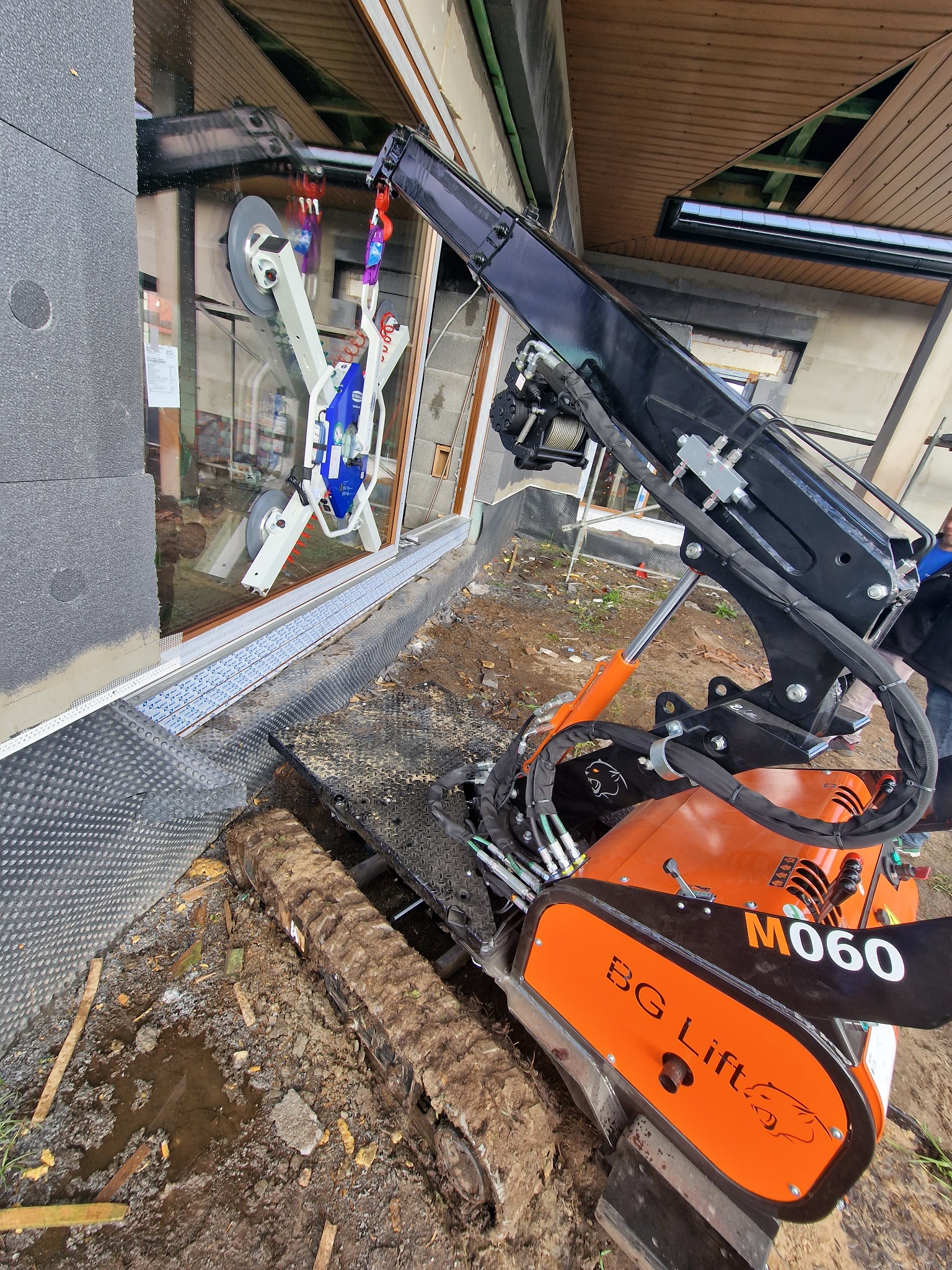 Pomarańczowy mini-dźwig gąsienicowy BG Lift M060 z przyssawkami, montujący dużą szybę w nowym budynku w trakcie budowy.