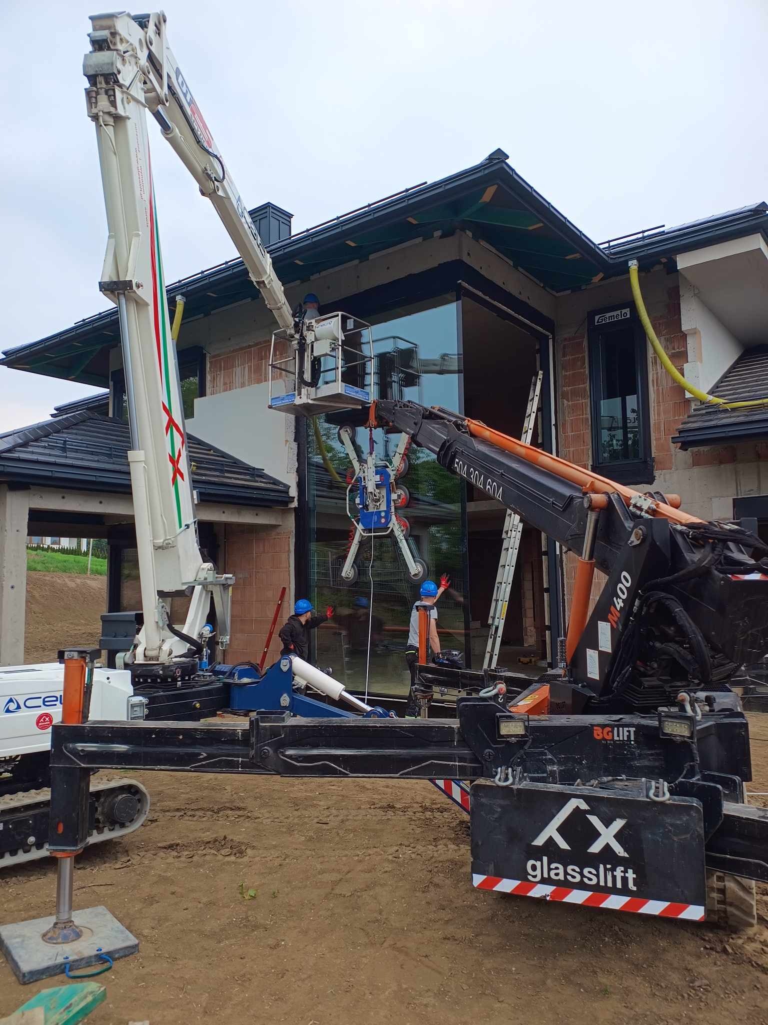 Montaż okna w domu jednorodzinnym przy użyciu podnośnika koszowego z przyssawkami. Dwóch pracowników w kaskach nadzoruje proces.