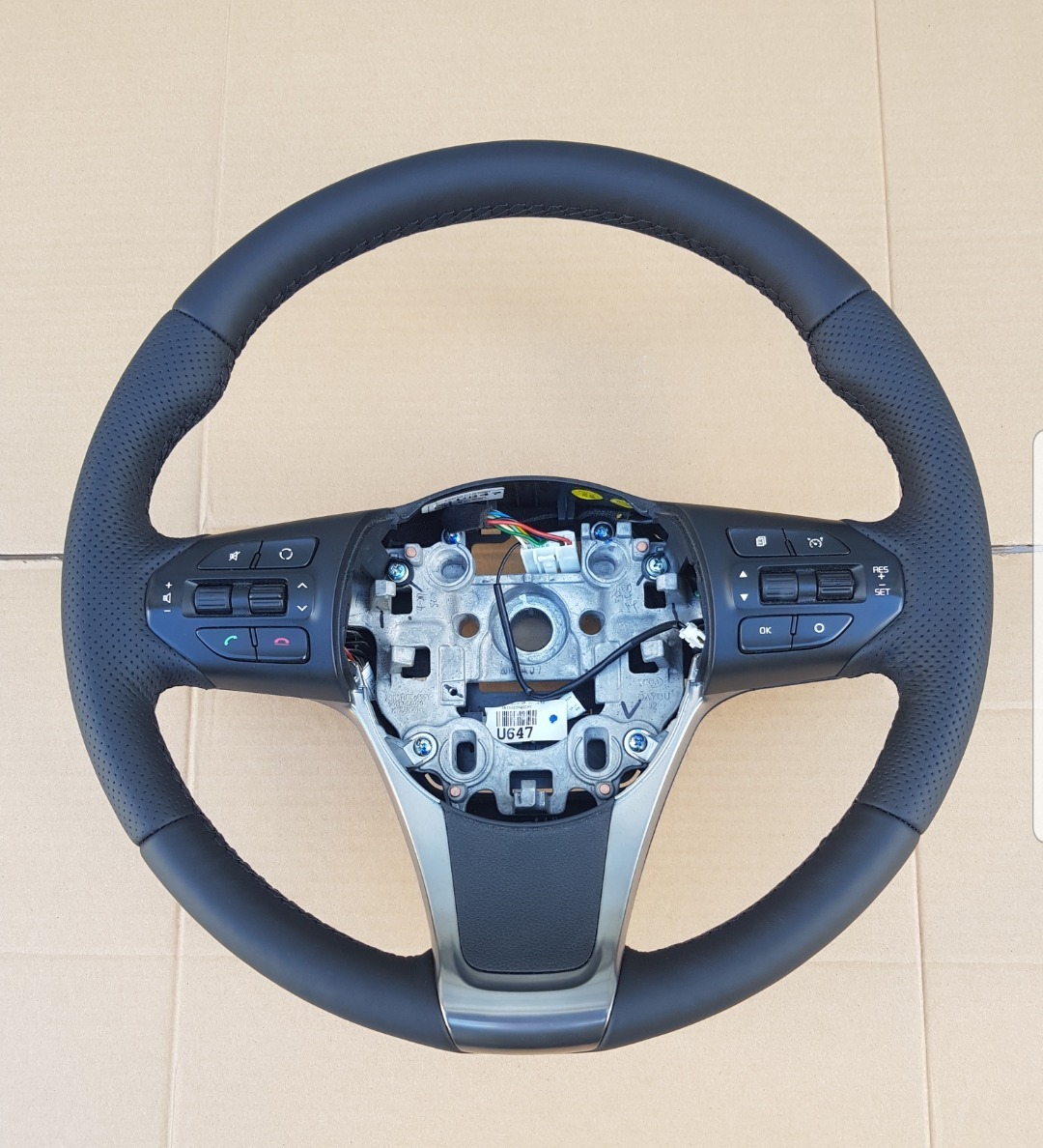 Obszyta skórą kierownica samochodowa z widocznymi elementami montażowymi i przyciskami funkcyjnymi, leżąca na kartonowym tle.