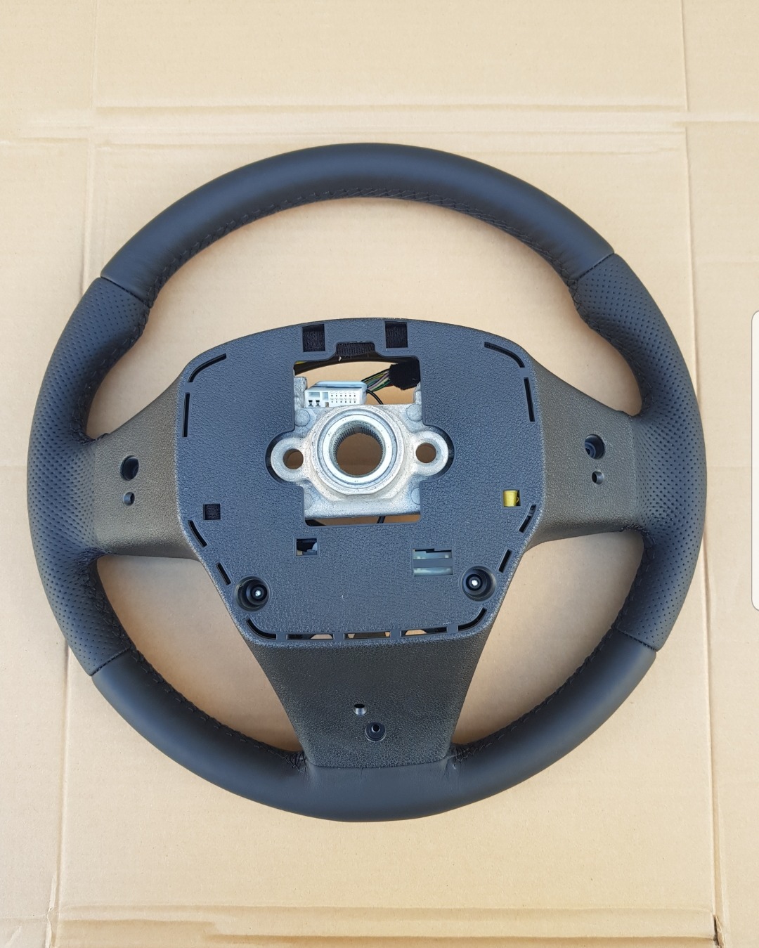 Obszyta czarną skórą kierownica samochodowa z perforowanymi bokami, widoczne mocowanie do kolumny kierowniczej, leżąca na kartonowym podłożu.
