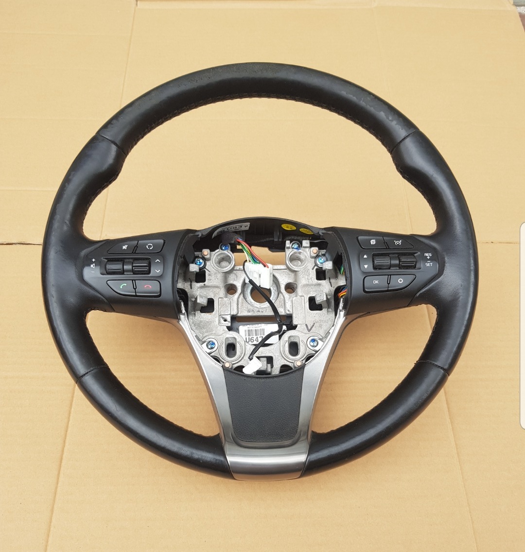 Używana kierownica samochodowa z widocznymi śladami eksploatacji na czarnej, skórzanej obręczy, z elementami sterowania i odsłoniętym mechanizmem mocowania na kartonowym tle.